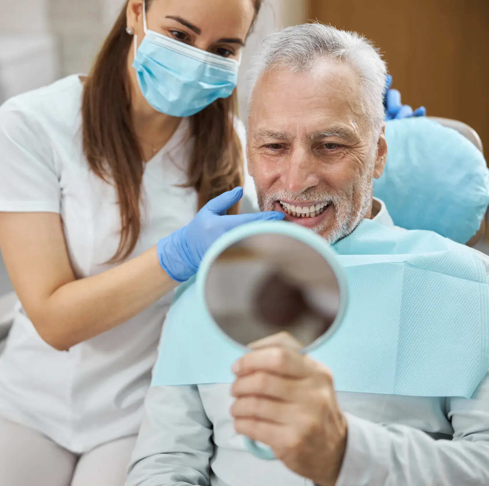 Smiling older man in dental chair checks his teeth in mirror while masked dentist assists him.