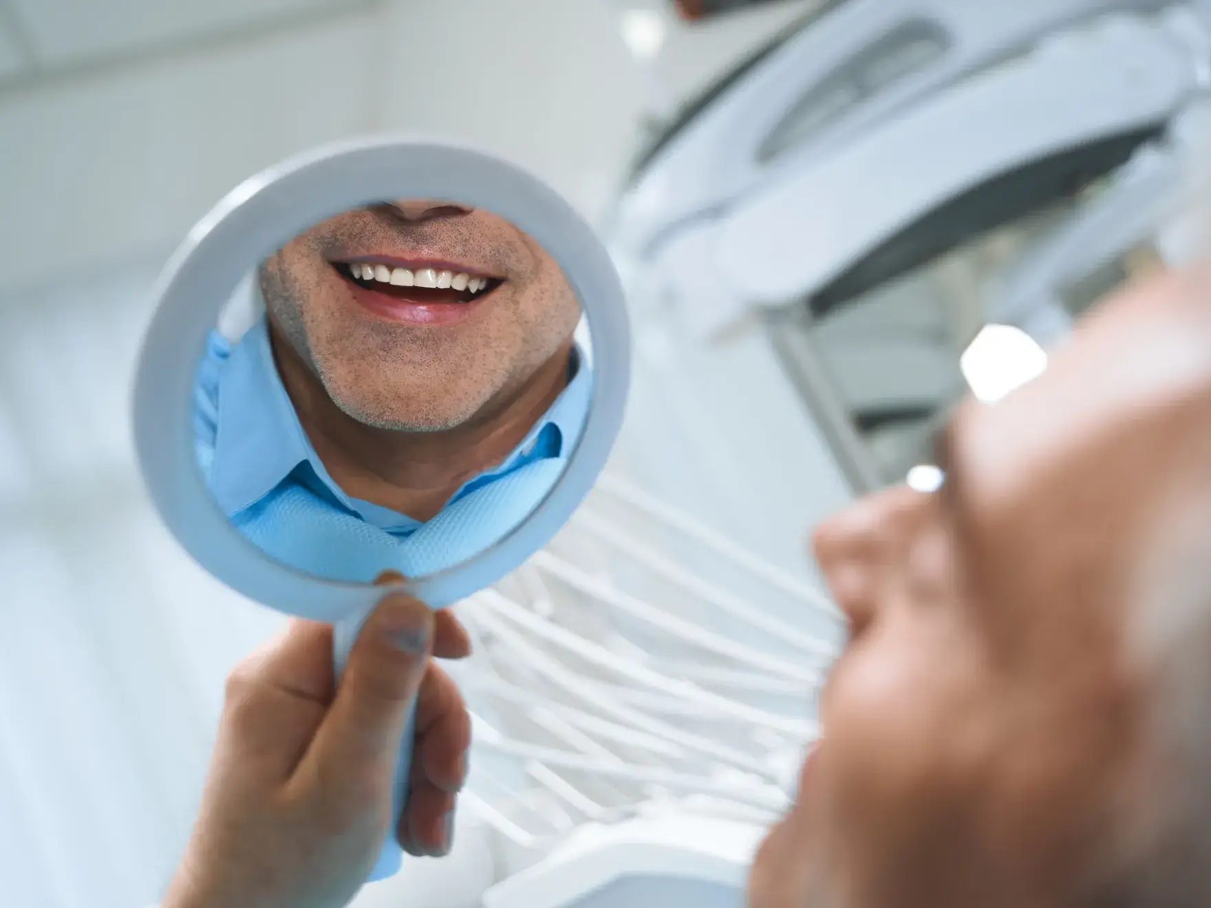 Man holding a small round mirror showing his smile and lower face close-up.