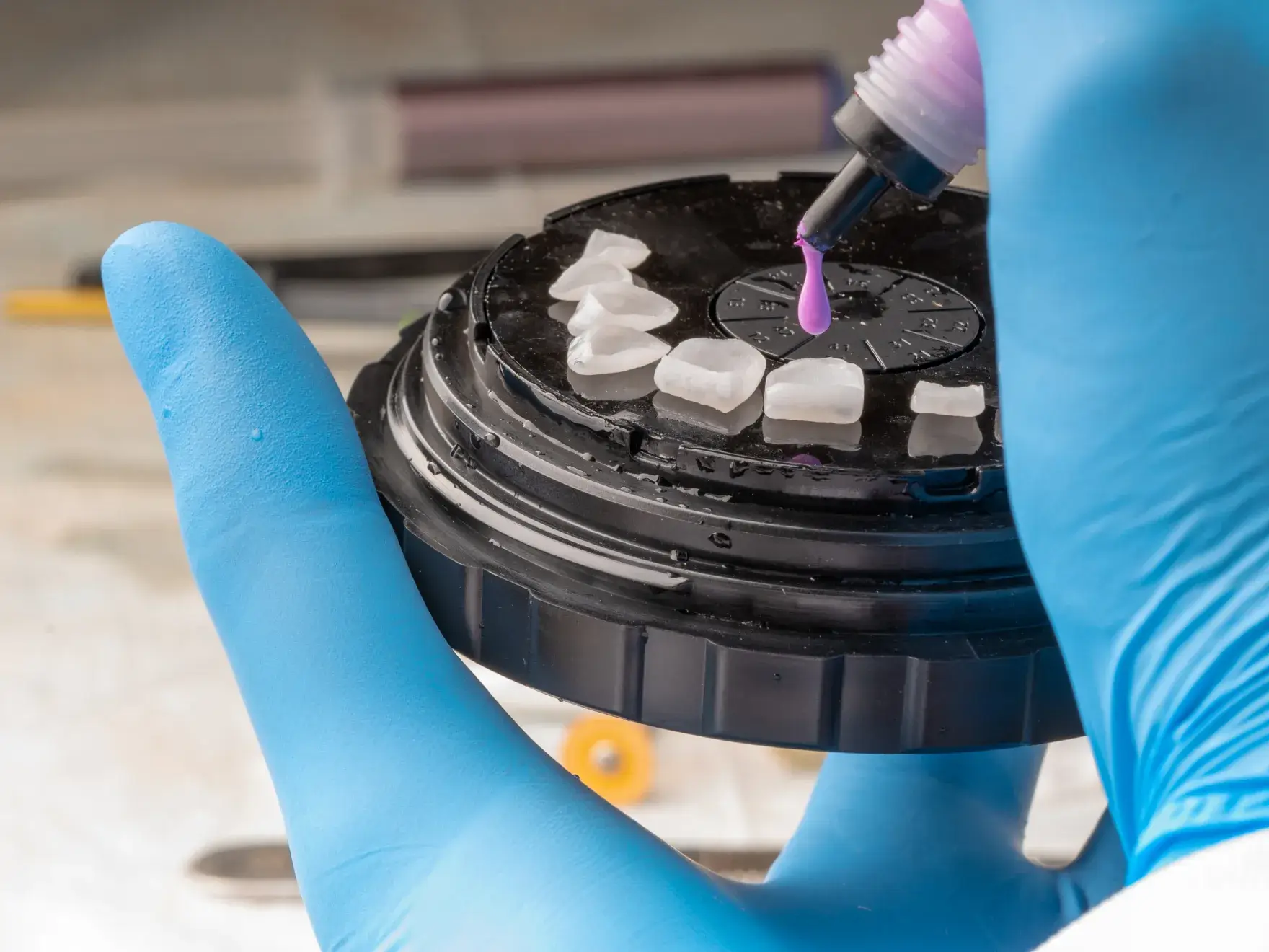 Gloved hand holding a dental mold as pink resin drops onto artificial teeth on a circular tray.
