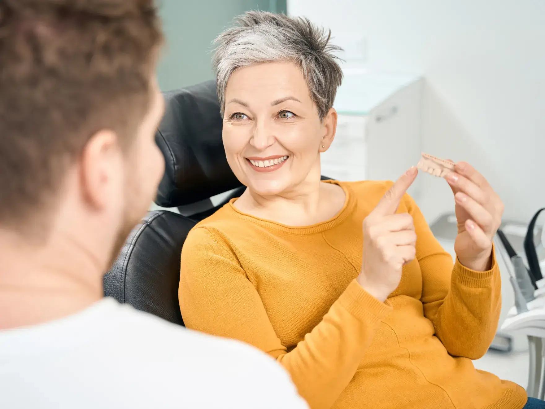 Smiling older woman in dental chair discussing dental model with dentist during consultation.