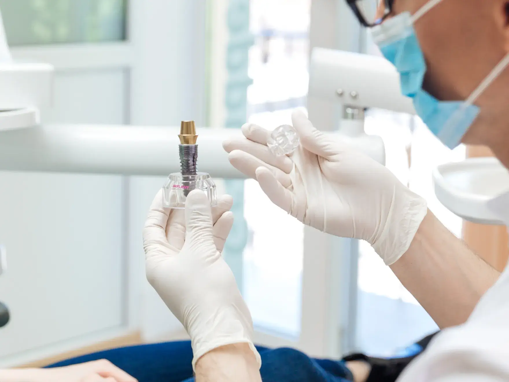 Dentist wearing mask and gloves holding a dental implant model and a clear mold in a clinic.