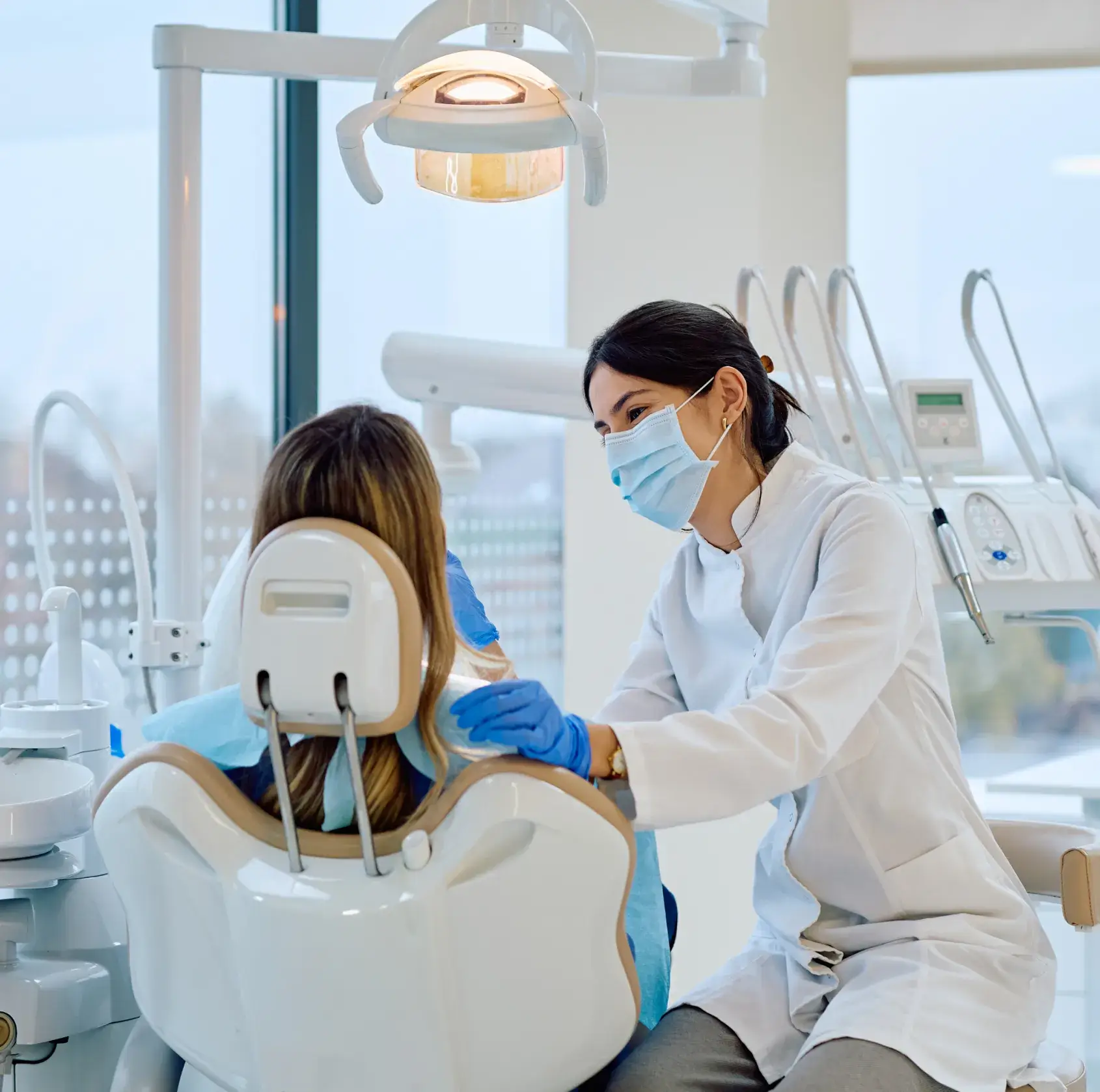 Dentist wearing mask and gloves comforting a patient seated in a dental chair.