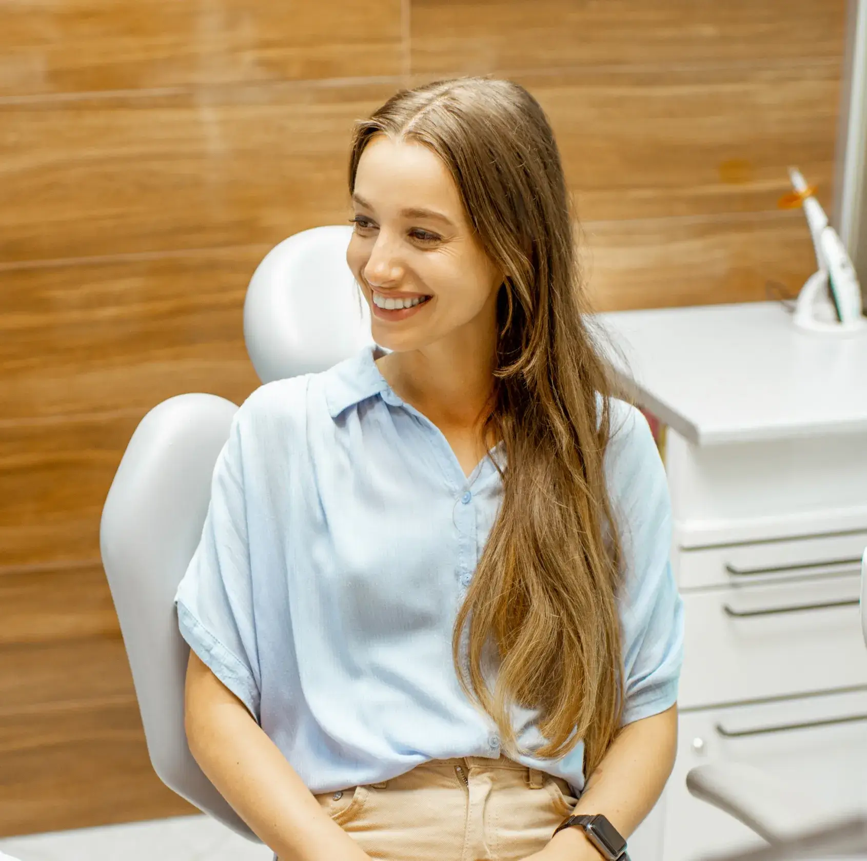 Smiling woman with long hair sits in a dental chair in a clinic room with wood panel walls.