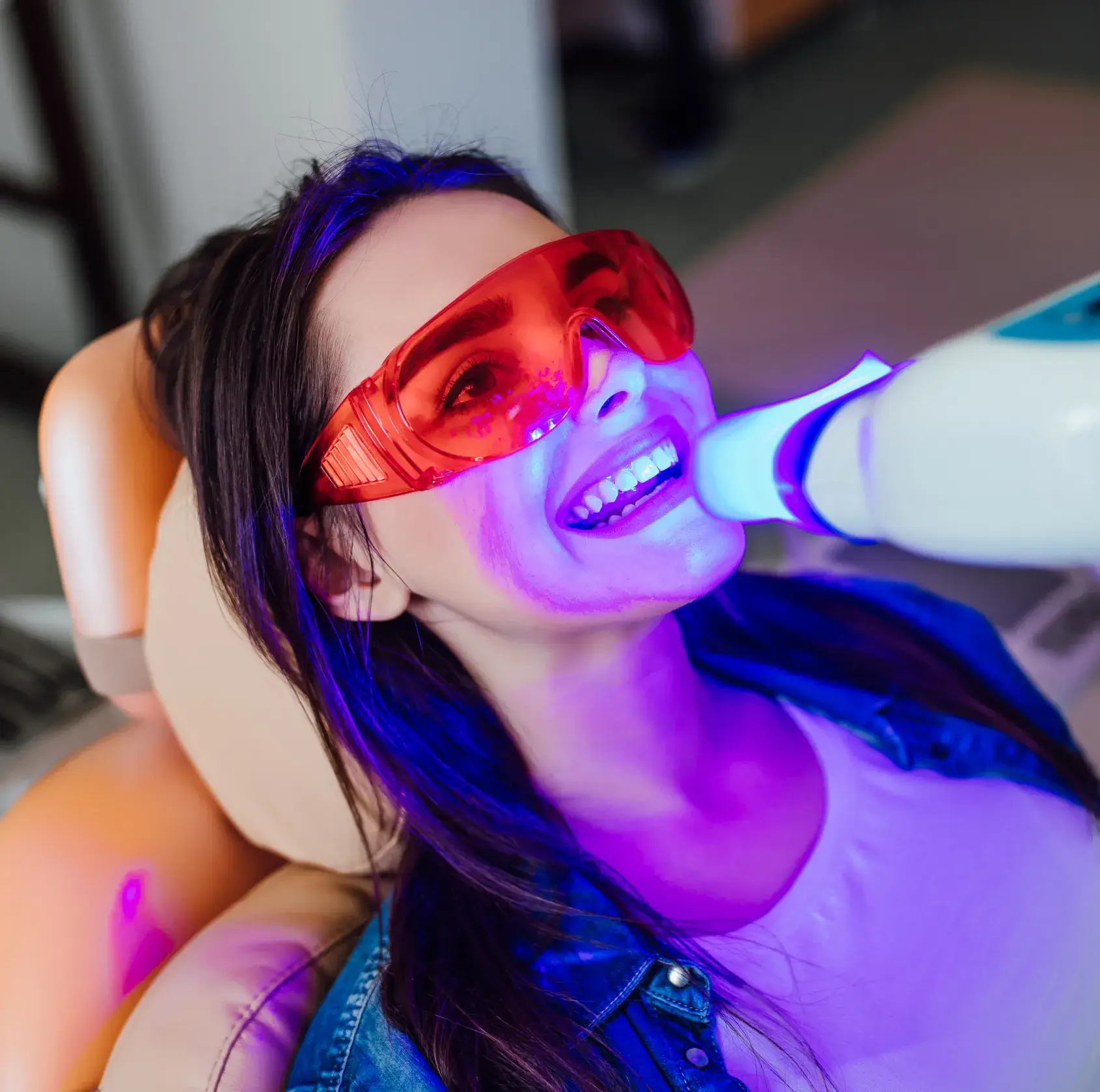 Woman wearing red protective glasses receiving blue light teeth whitening treatment reclined in chair.