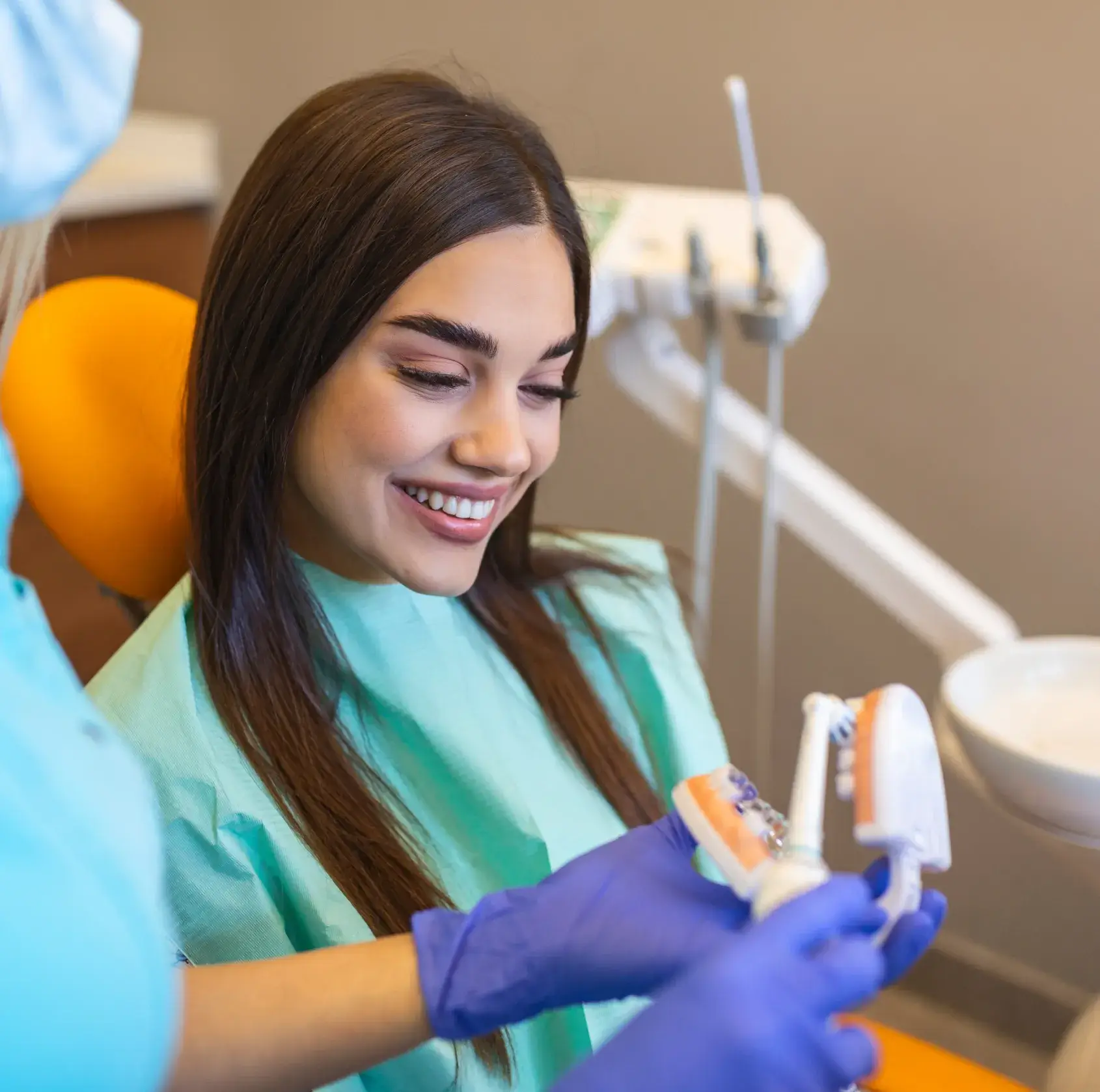 Dentist explains oral care using a dental model to a smiling patient in a clinic chair.