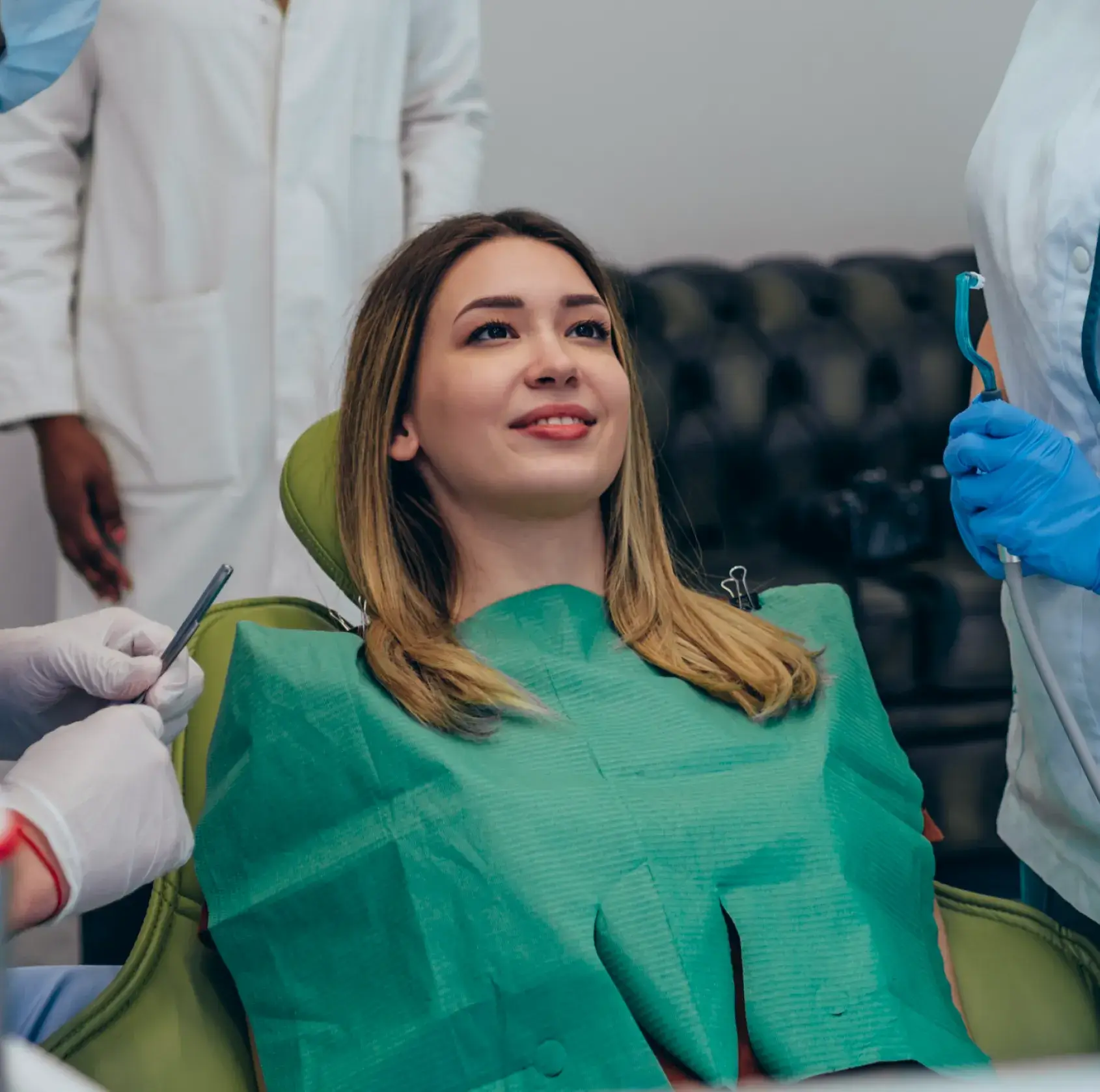 Smiling woman reclining in a dental chair with green bib surrounded by dental professionals.