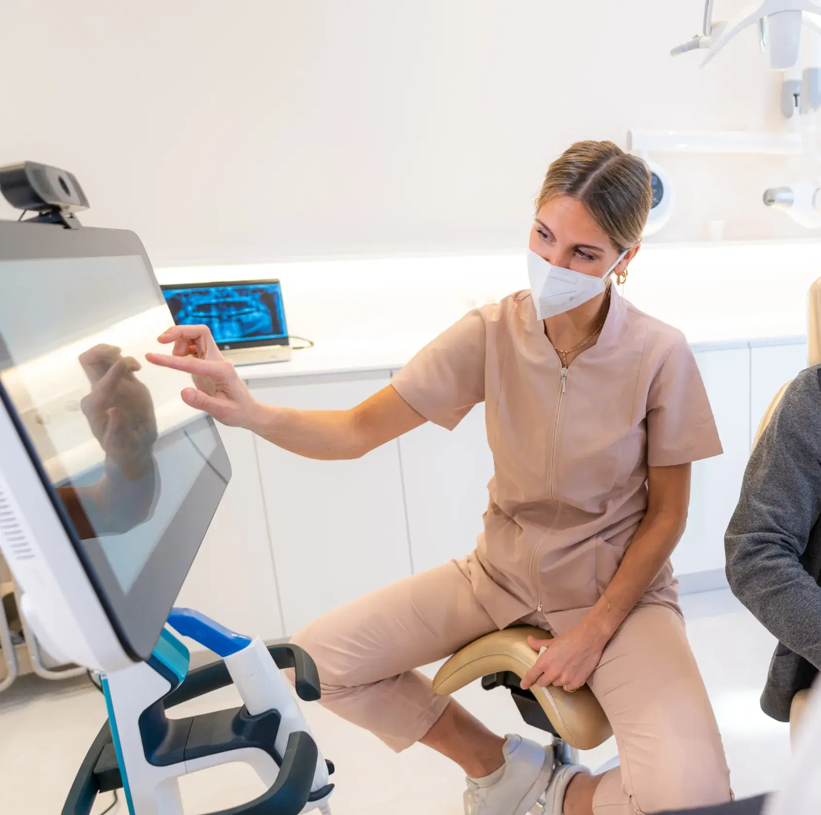 Dentist in scrubs and mask pointing at screen in a dental office, showing information to a patient.
