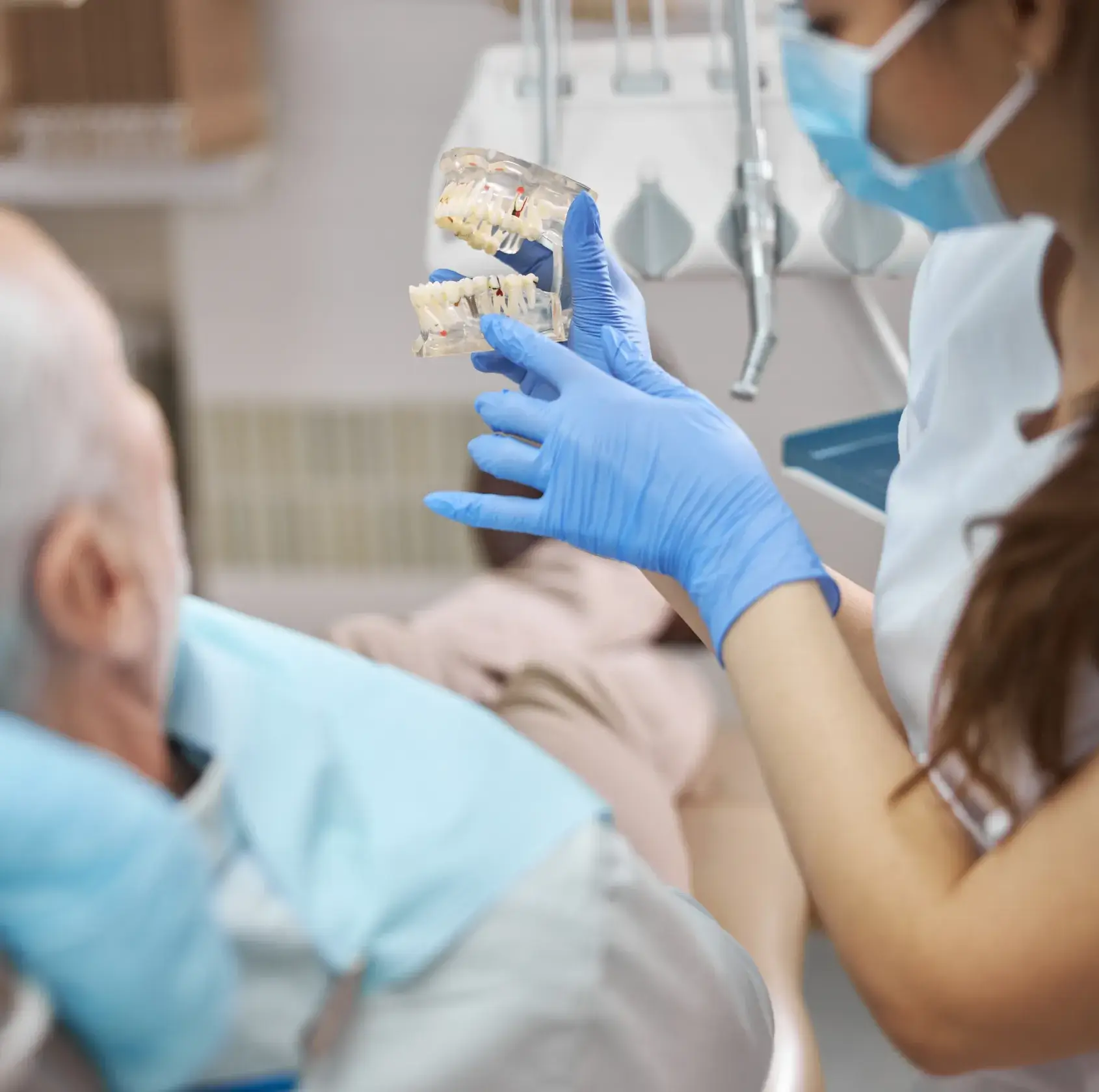 Dentist wearing blue gloves holds a dental model explaining dental implants to an elderly patient.