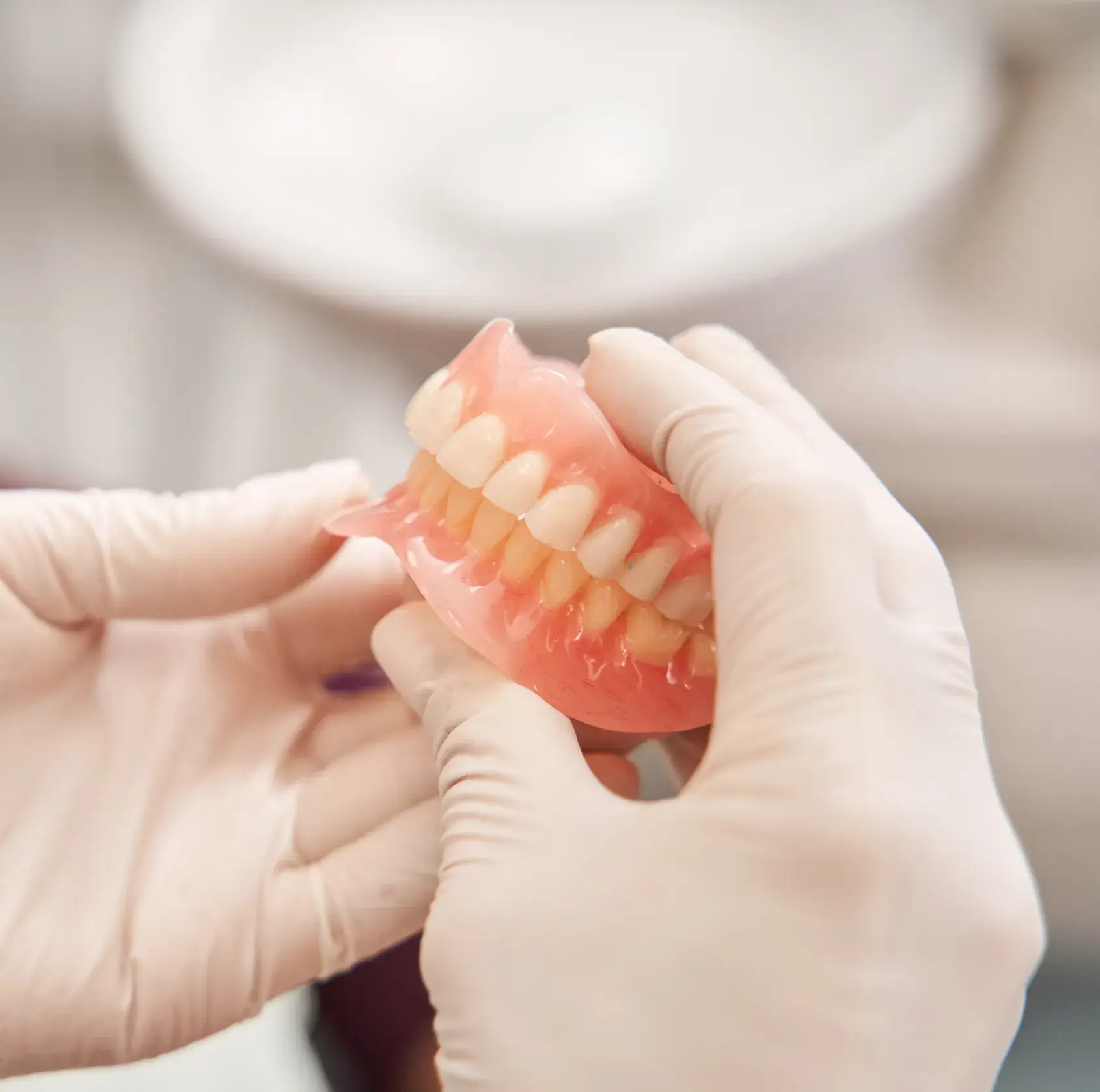 Hands in gloves holding a set of pink dentures with white artificial teeth.