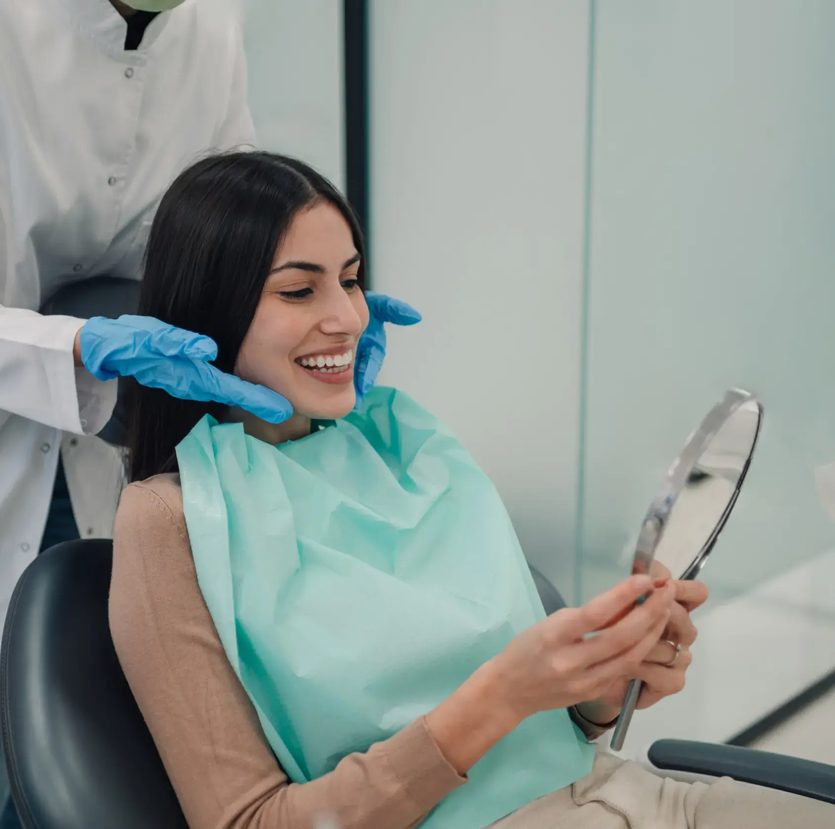 Woman with dark hair smiling in a dental chair holding a mirror, dentist's hands in blue gloves near her face.