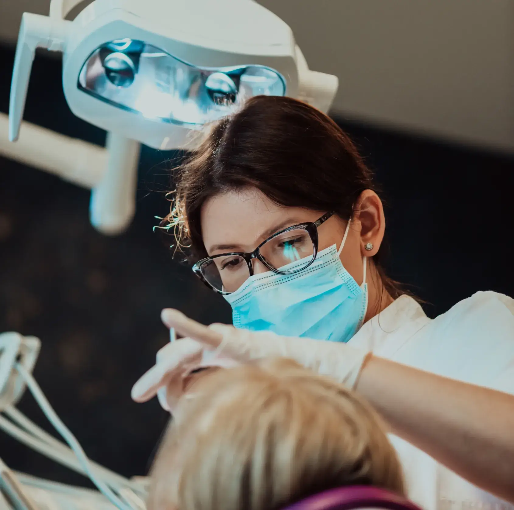 Female dentist wearing glasses and mask working on a patient under a dental light.