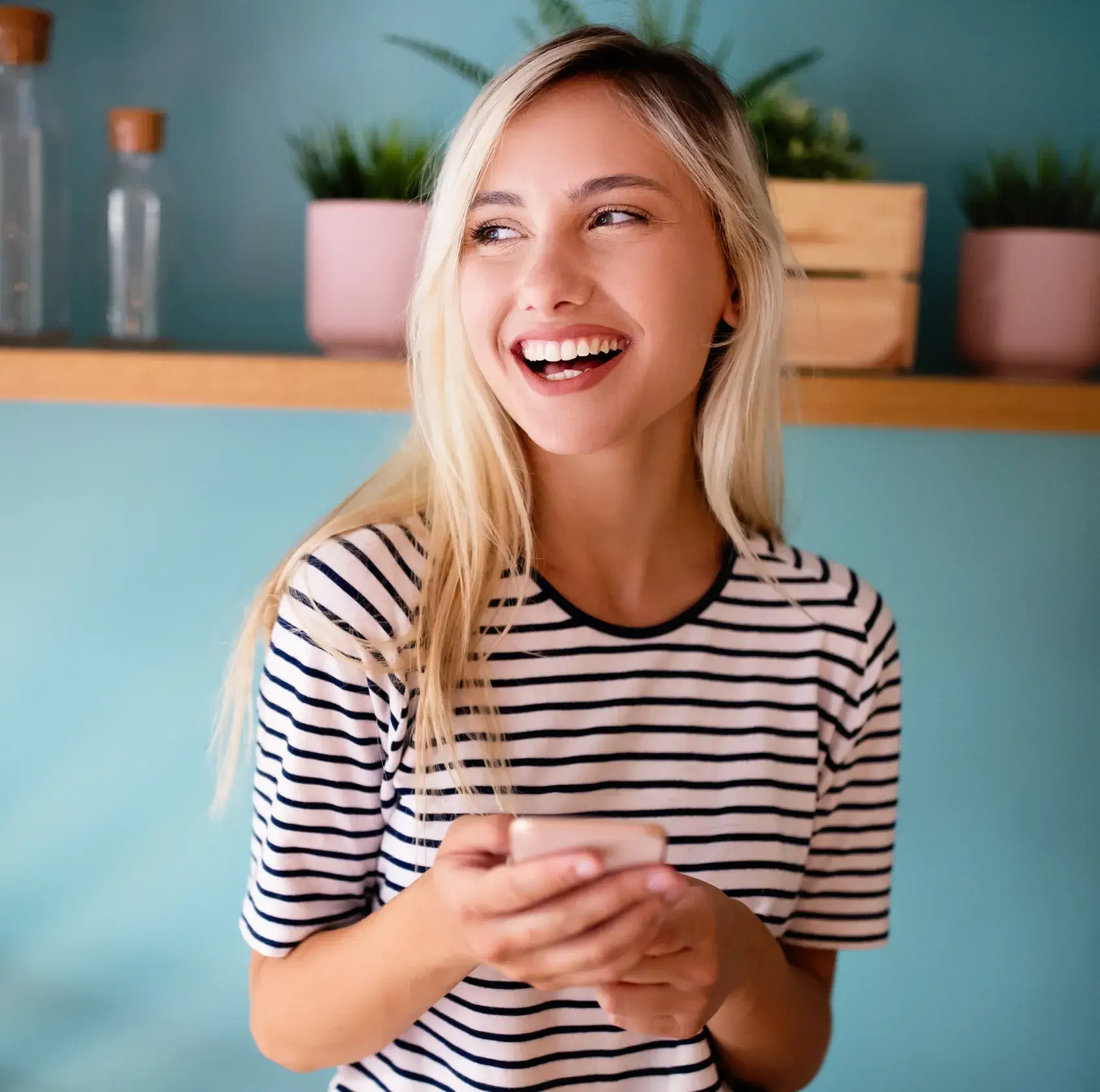 Smiling blonde woman in striped shirt holding a phone with a shelf and plants in the background.
