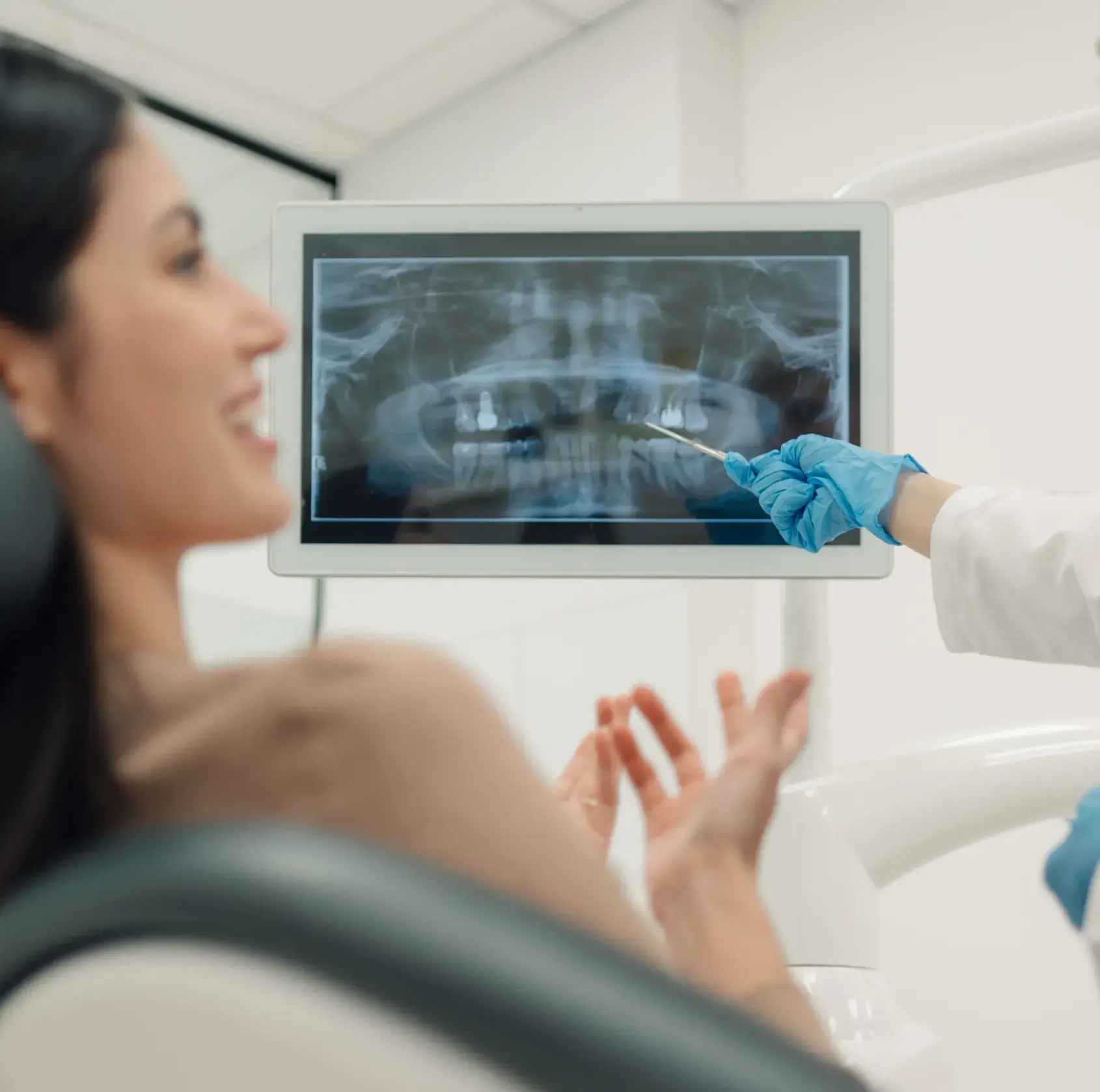 Dentist wearing blue gloves pointing to a dental X-ray on a screen while patient looks on smiling.