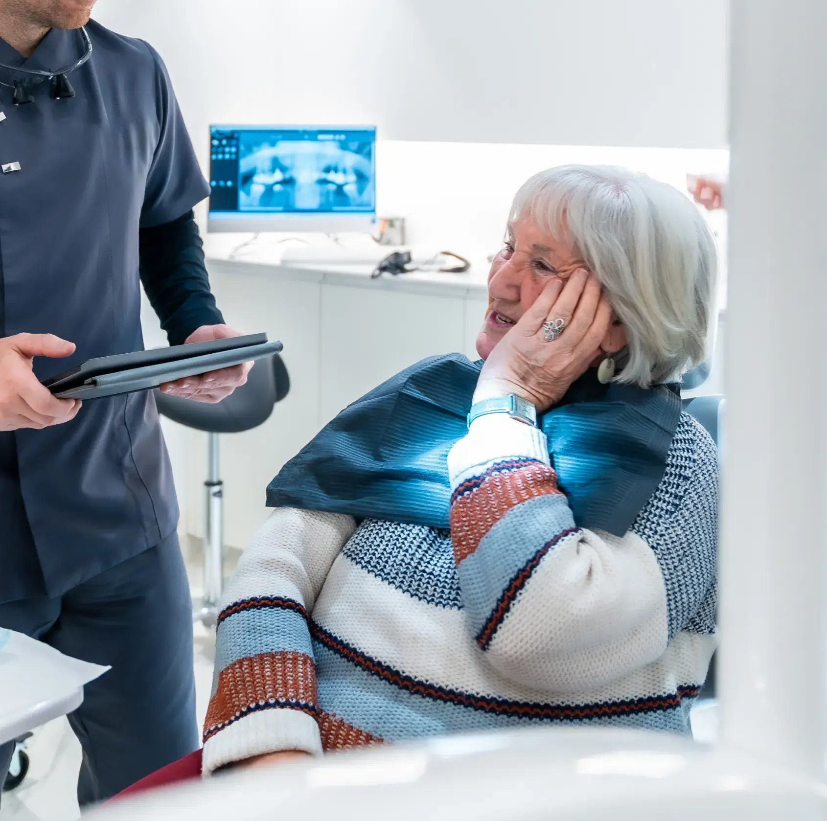 Elderly woman holding her cheek talking to a dentist in a dental office with X-ray on screen.