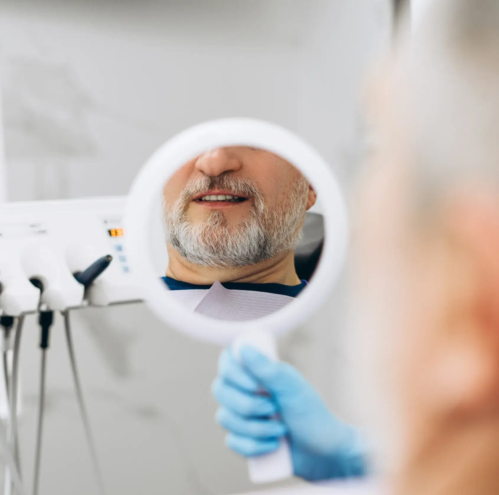 Middle-aged man with gray beard smiles looking at his teeth in a dental mirror held by a gloved hand.