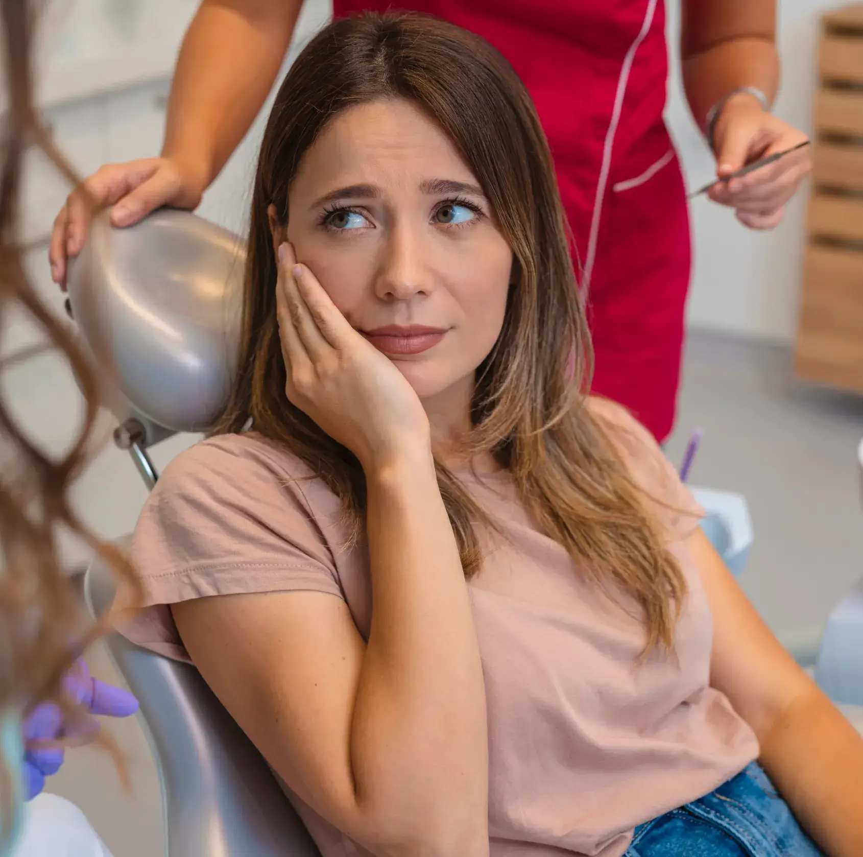 Woman with tooth pain sitting in a dentist's chair, holding her cheek with a worried expression.