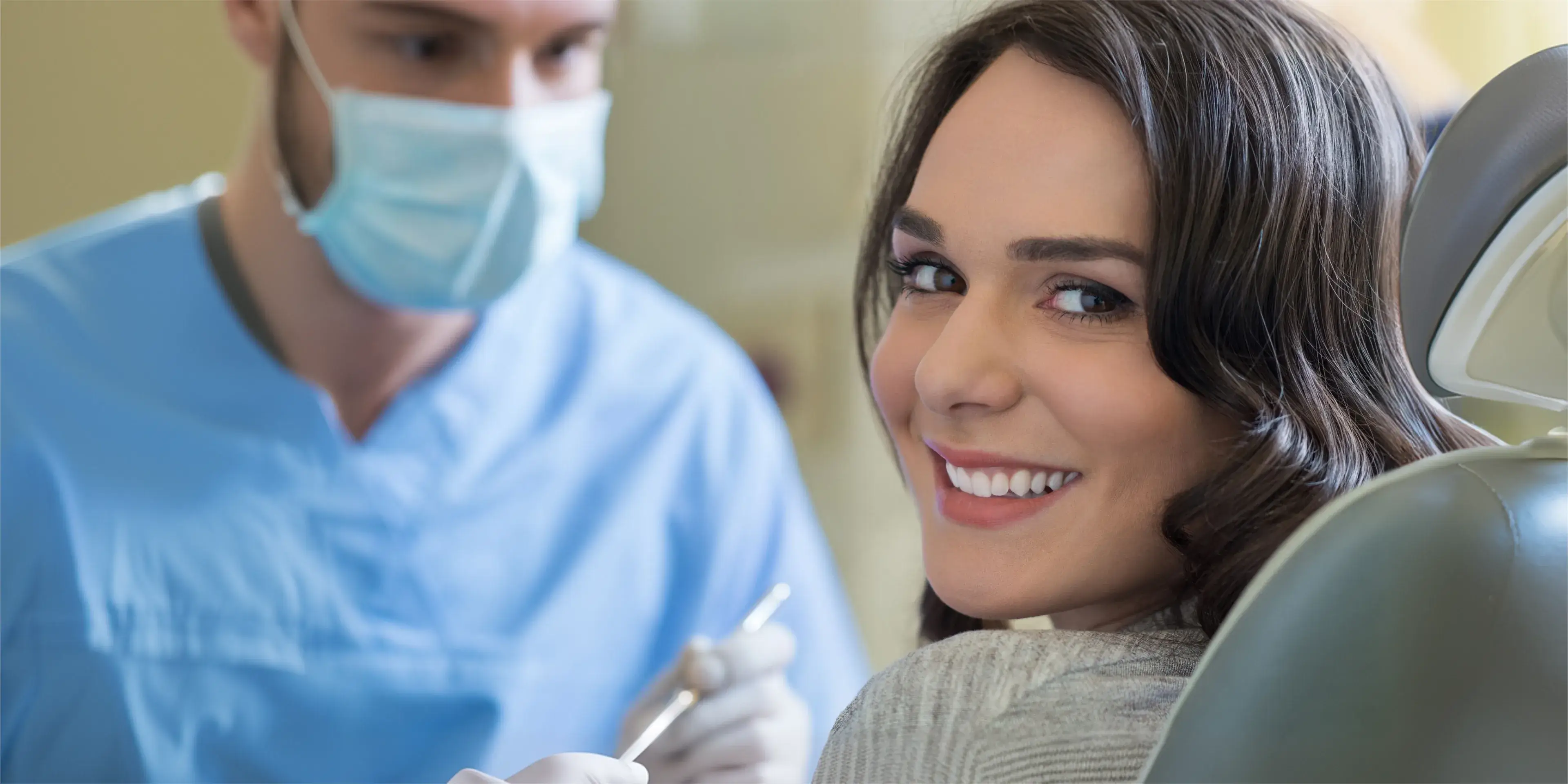 Smiling woman sitting in dental chair with dentist in mask and blue scrubs behind her.