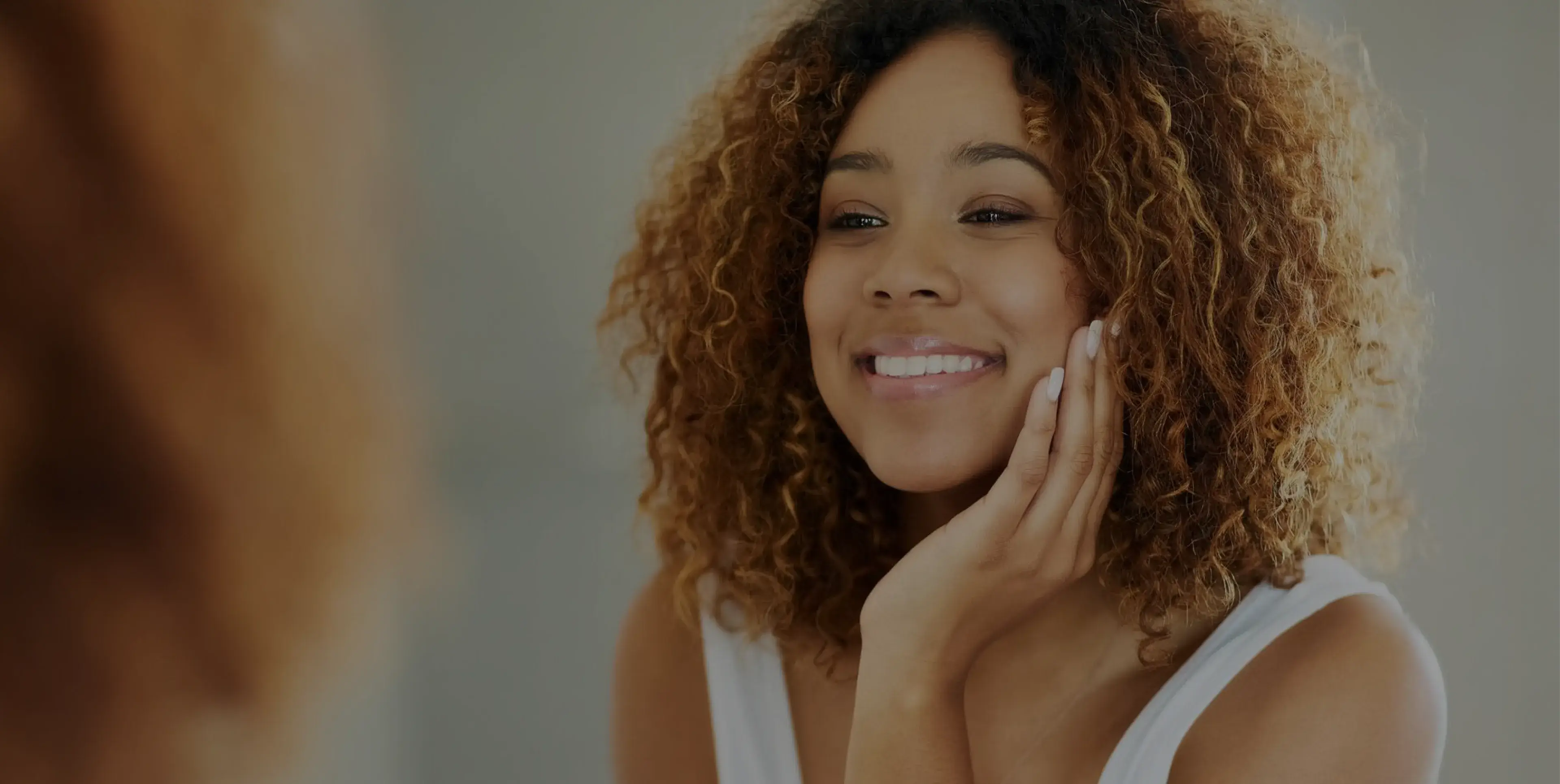 Smiling woman with curly hair touching her cheek looking at herself in the mirror.