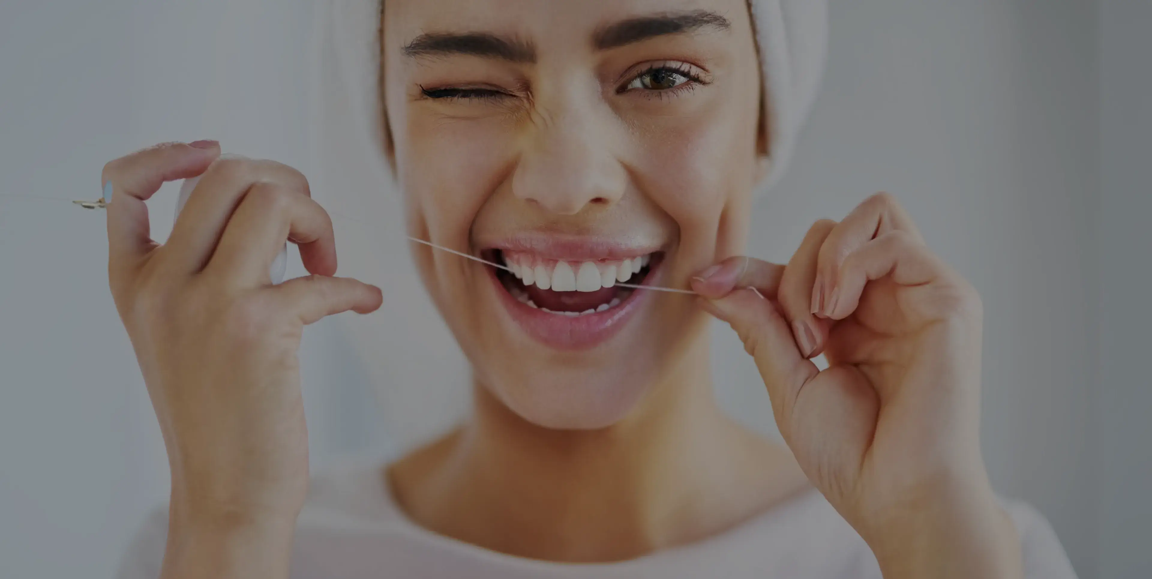 Smiling woman with towel on head flossing her teeth and winking with one eye.