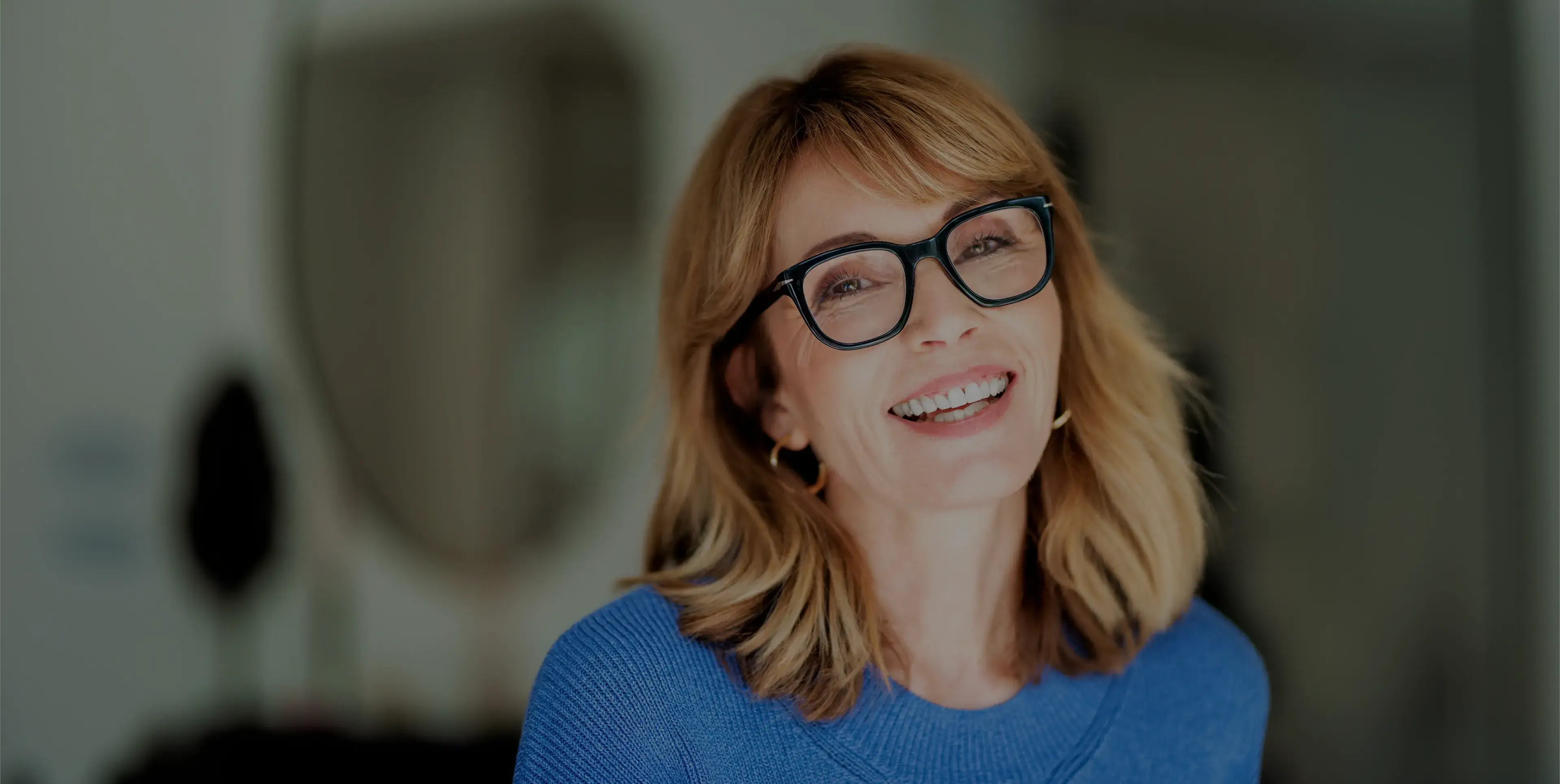 Smiling middle-aged woman with blonde hair and black glasses wearing a blue sweater indoors.