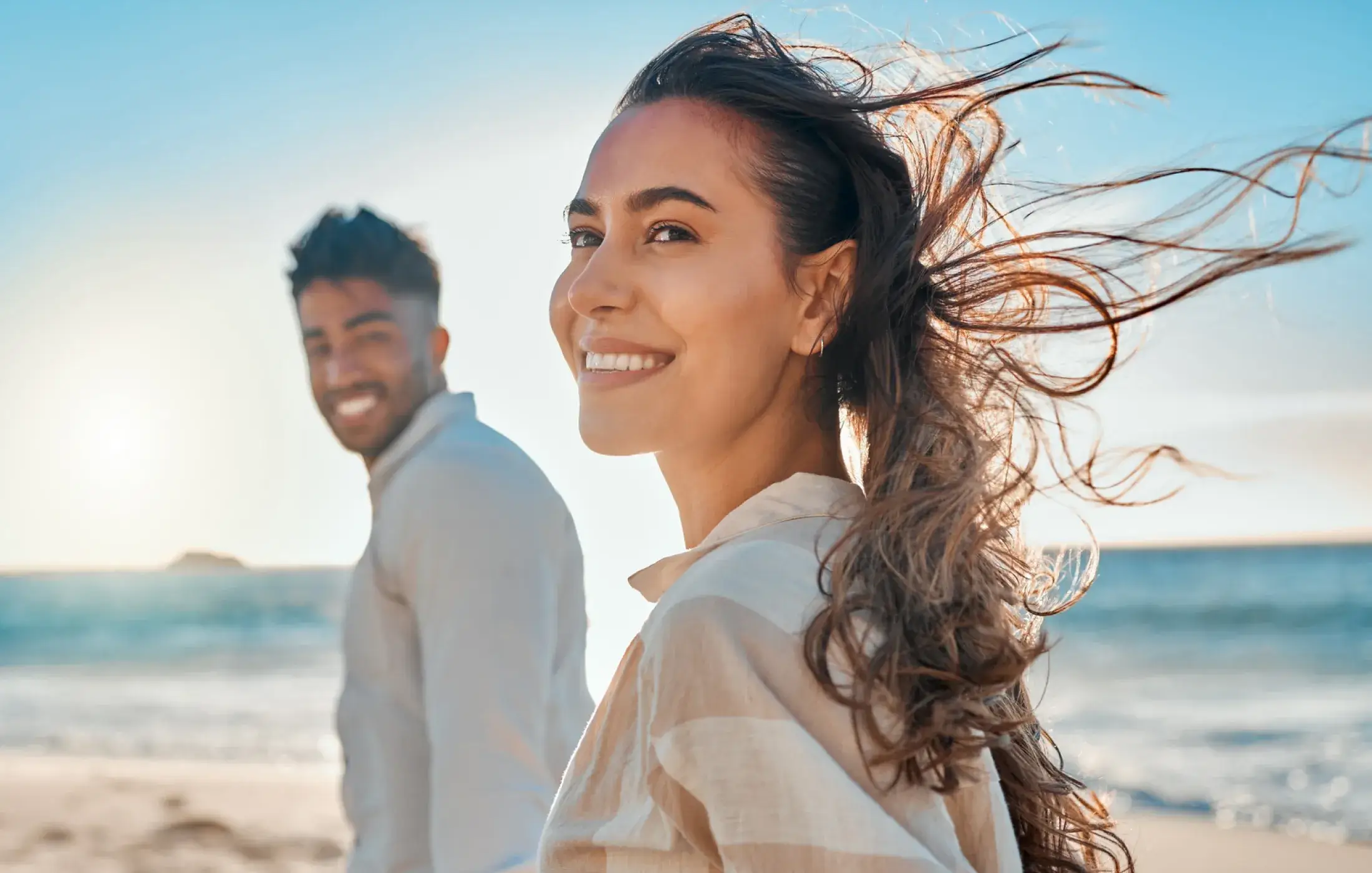Smiling woman with windblown hair and a man in the background on a sunny beach.