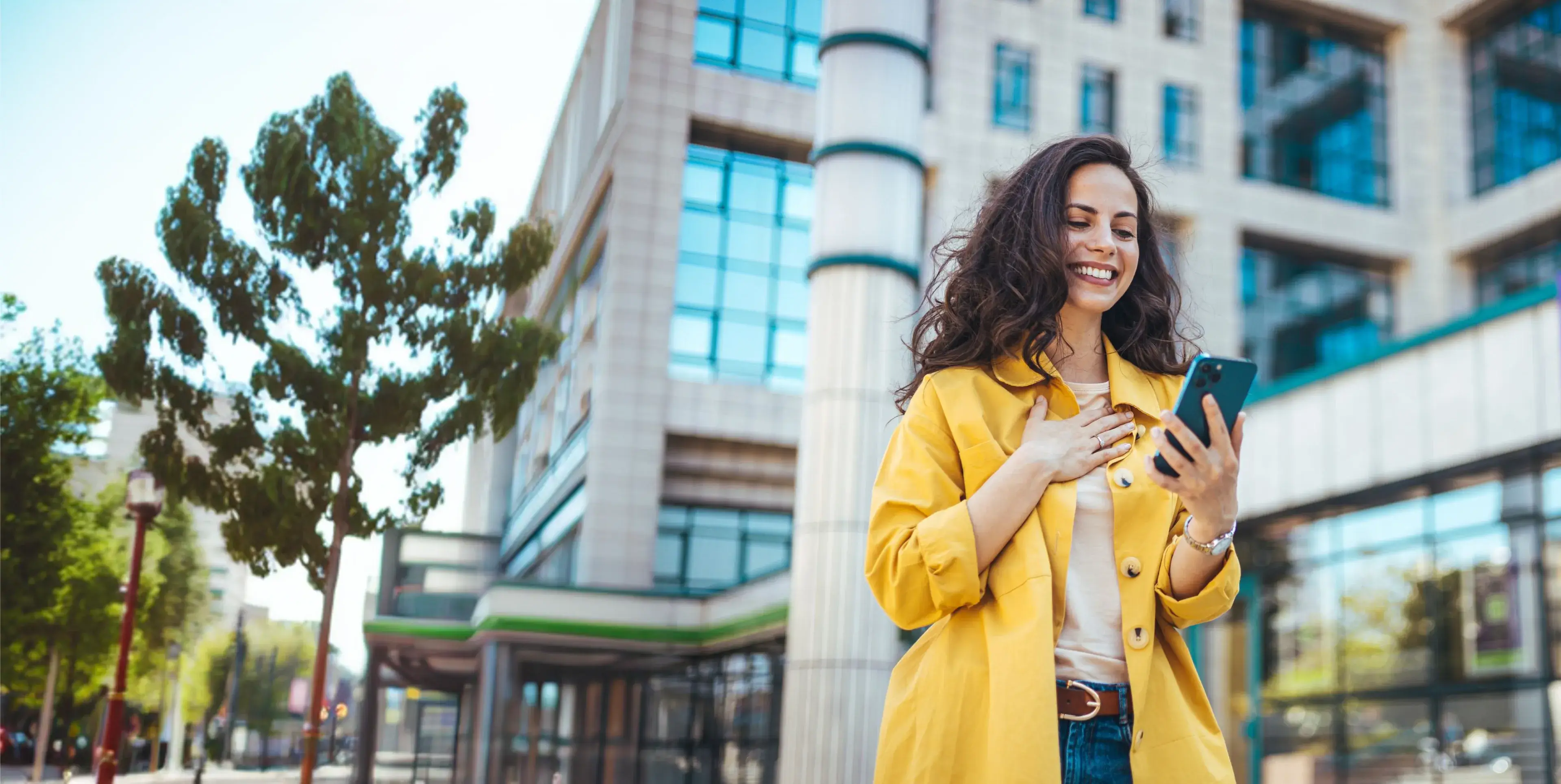 Smiling woman in yellow jacket looks at smartphone outside near modern building on sunny day.
