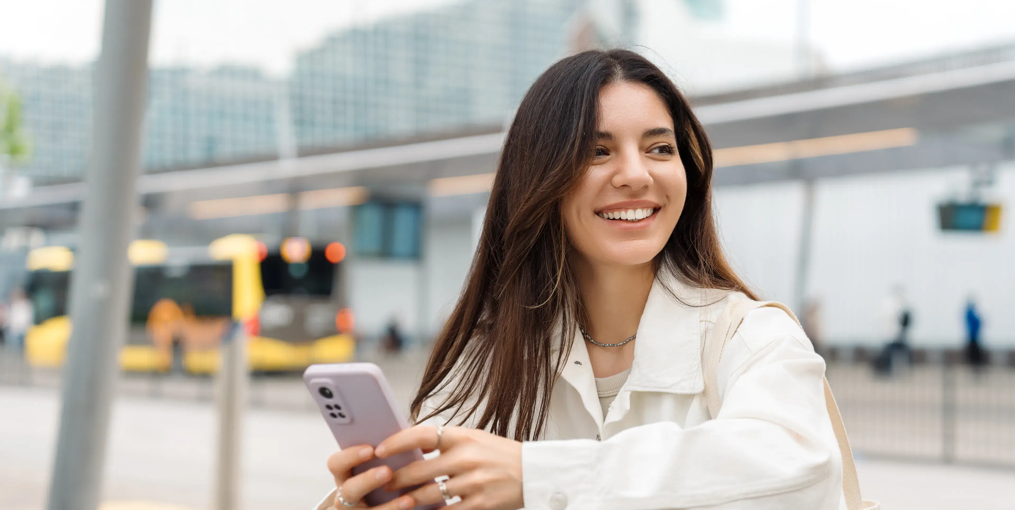 Smiling woman holding smartphone outdoors near bus station with blurred yellow buses in background.