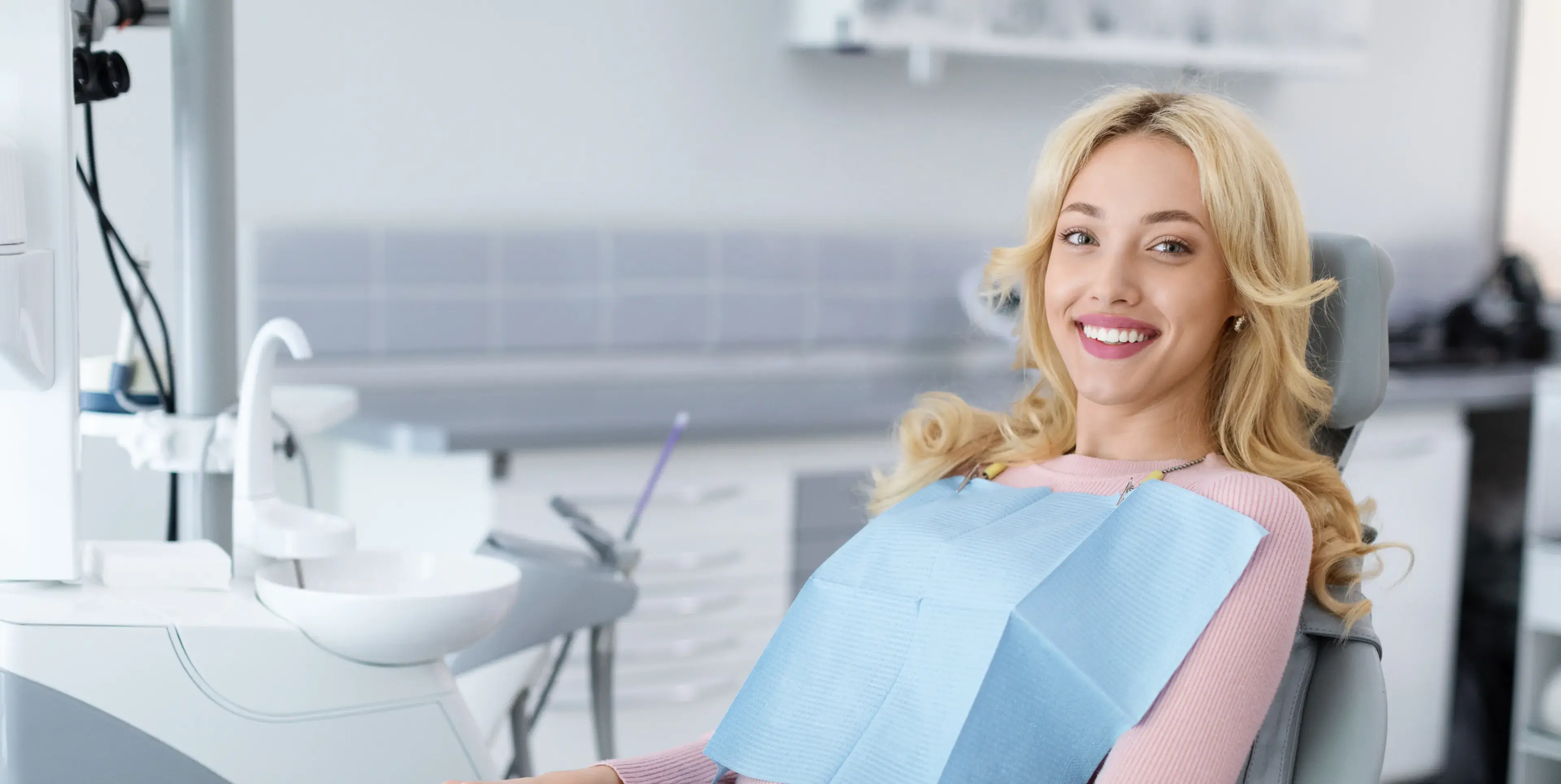 Smiling young woman with blonde hair sitting in a dental chair wearing a blue dental bib.