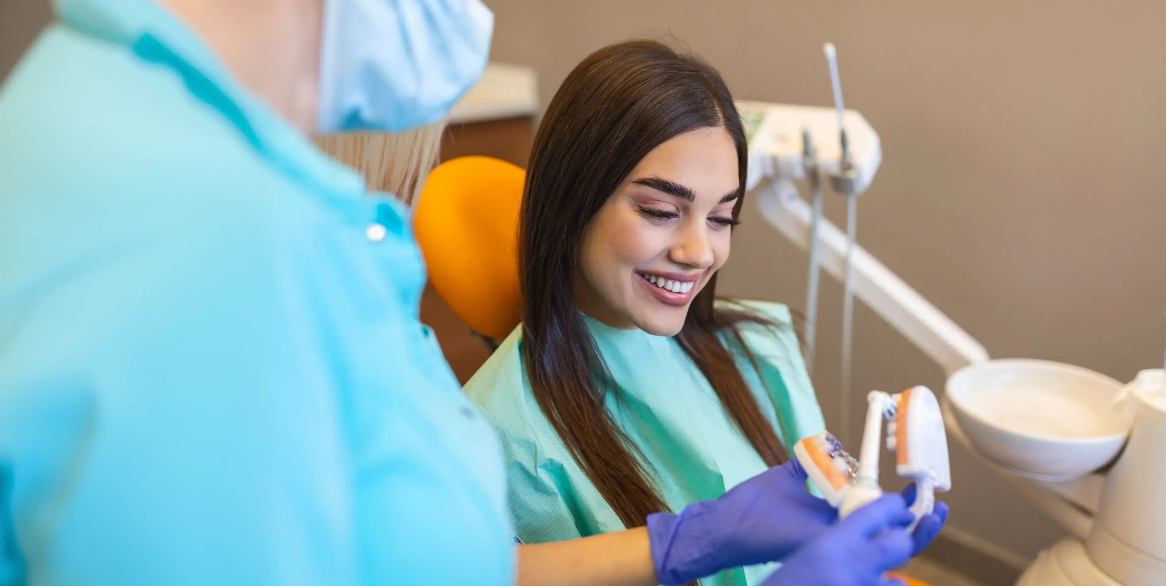 Smiling woman in dental chair watching dentist demonstrate dental hygiene with model teeth.