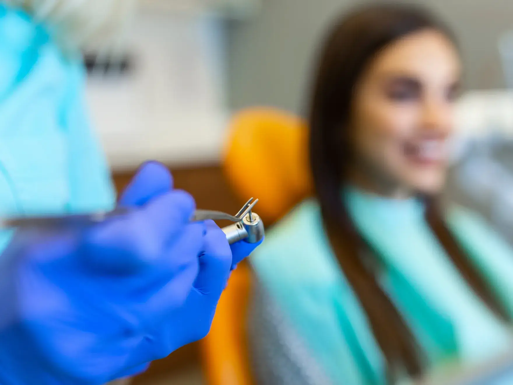 Dentist wearing blue gloves holding a dental drill near a smiling patient in a dental chair.