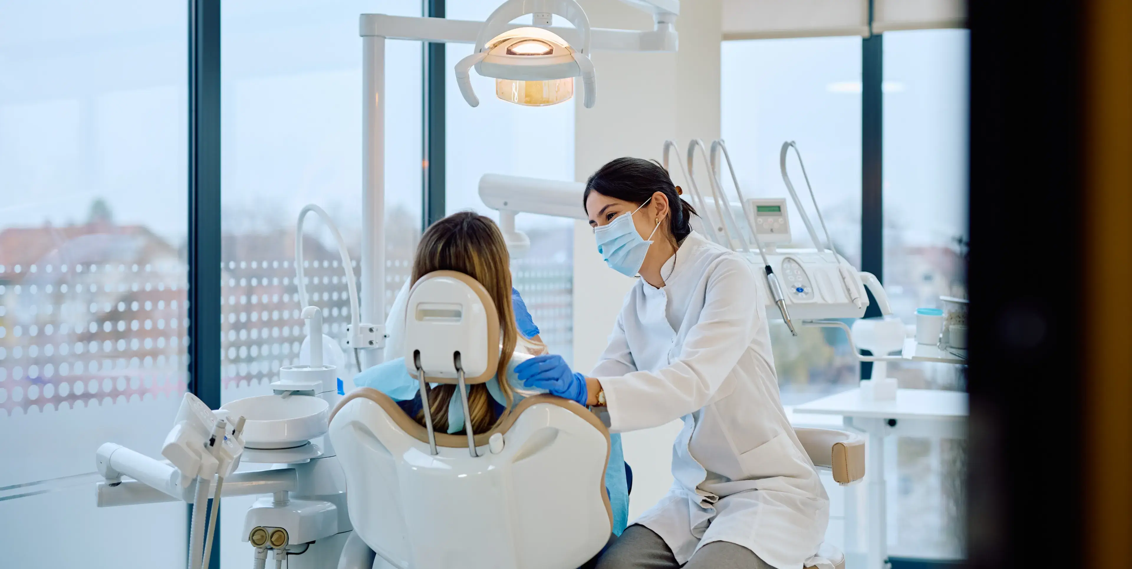Dentist in mask and gloves talking to a patient sitting in a dental chair in a bright clinic.