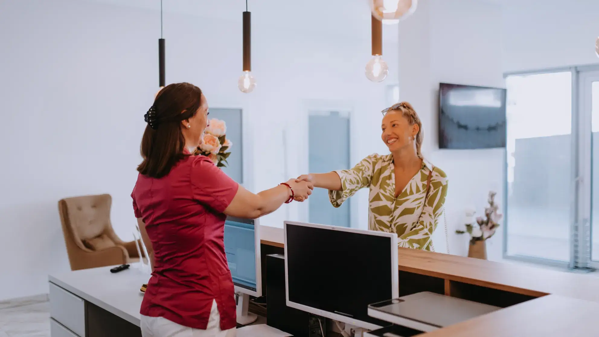Woman in red scrubs shaking hands with smiling woman in a green and white blouse at a reception desk.