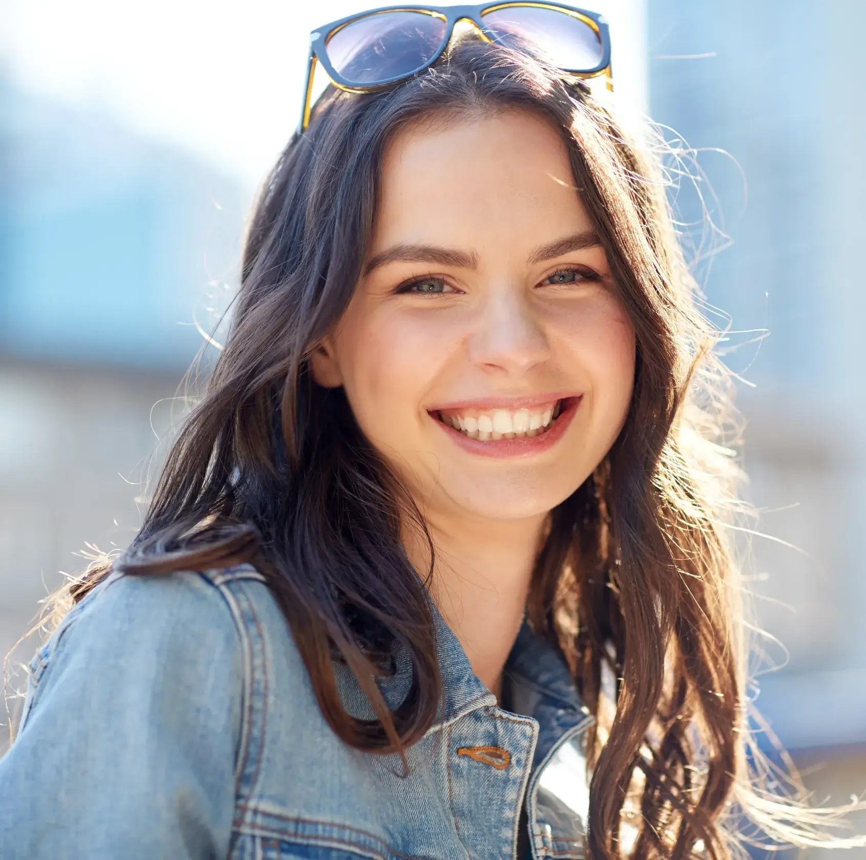Smiling young woman with long dark hair wearing a denim jacket and sunglasses on her head outdoors.