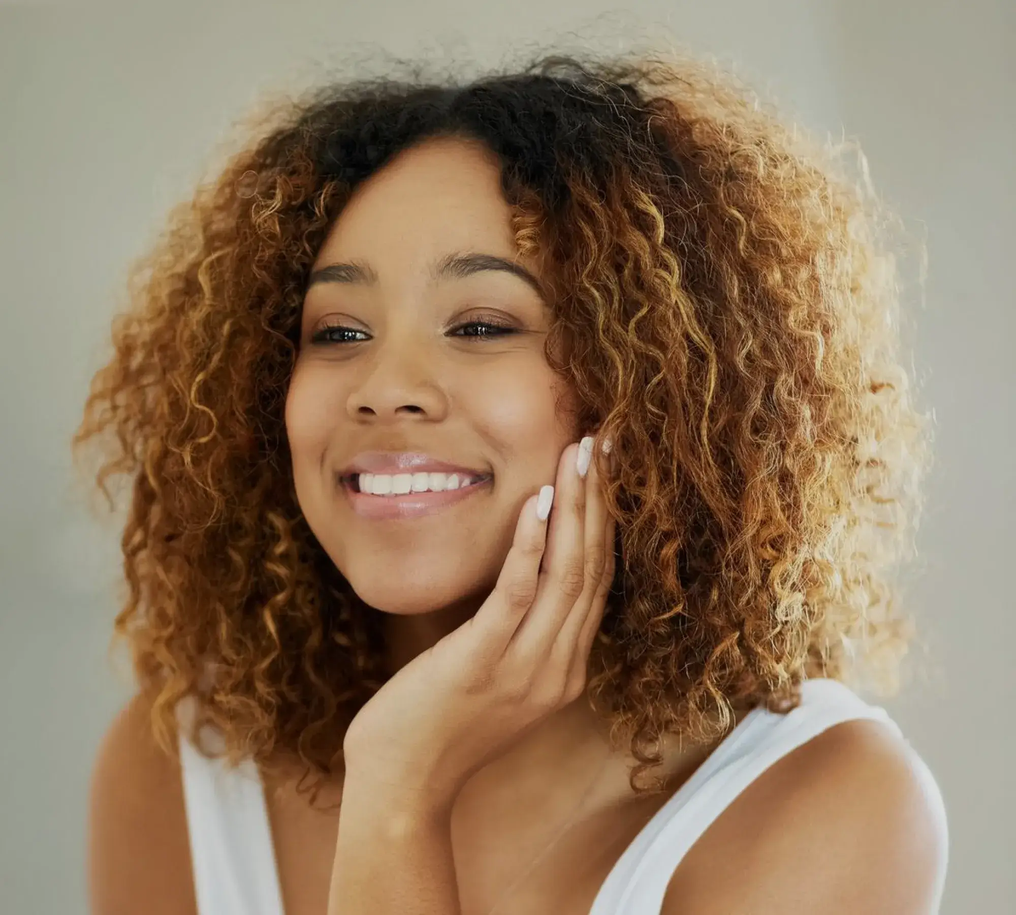 Smiling woman with curly hair resting her face on her hand against a neutral background.