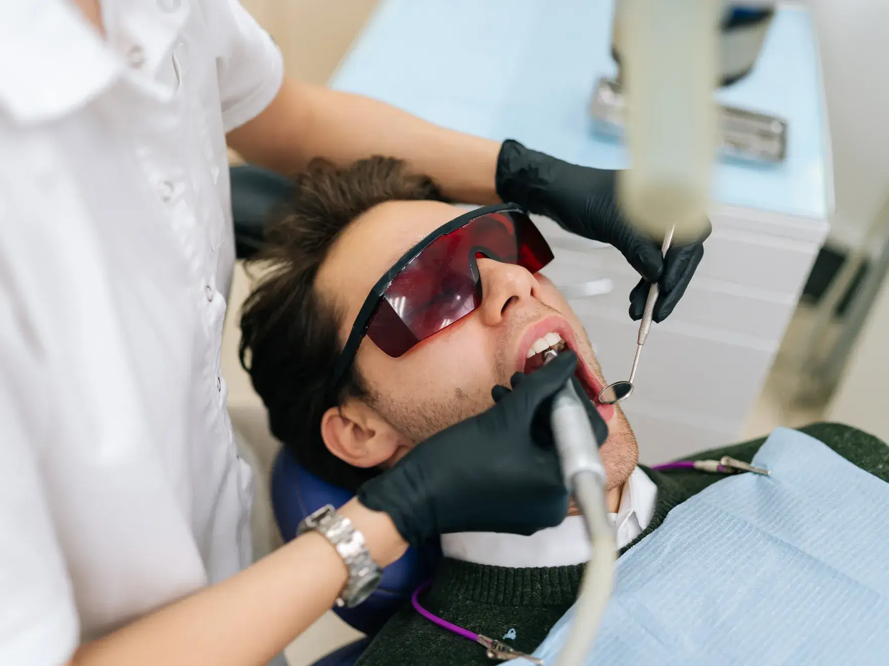 Dentist wearing black gloves treating a male patient wearing protective red glasses.