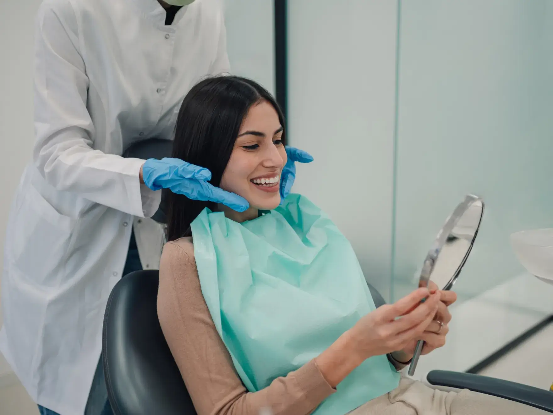 Dentist with blue gloves examines smiling woman in dental chair holding a hand mirror.
