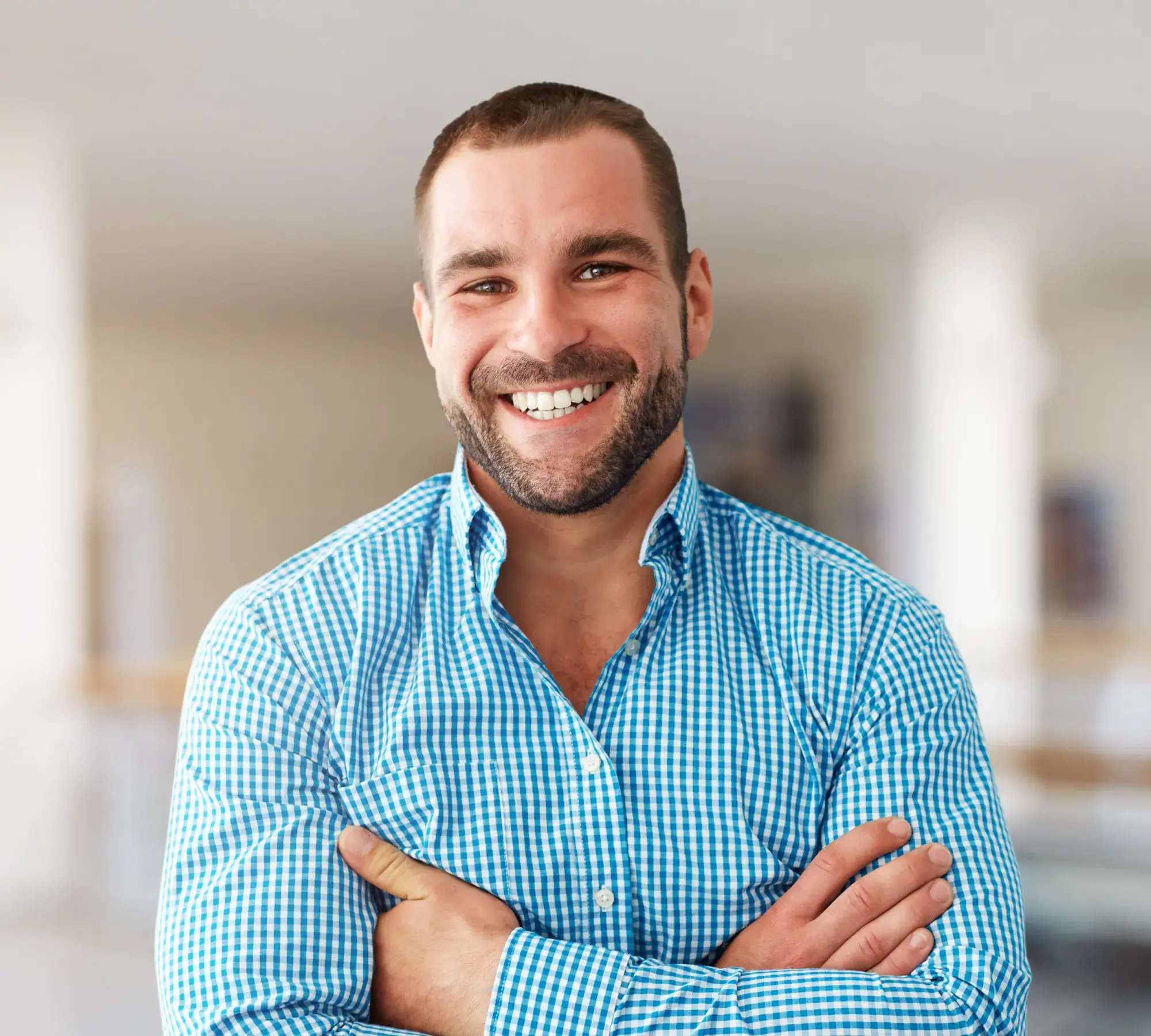 Smiling man with beard wearing blue checkered shirt stands with arms crossed indoors.