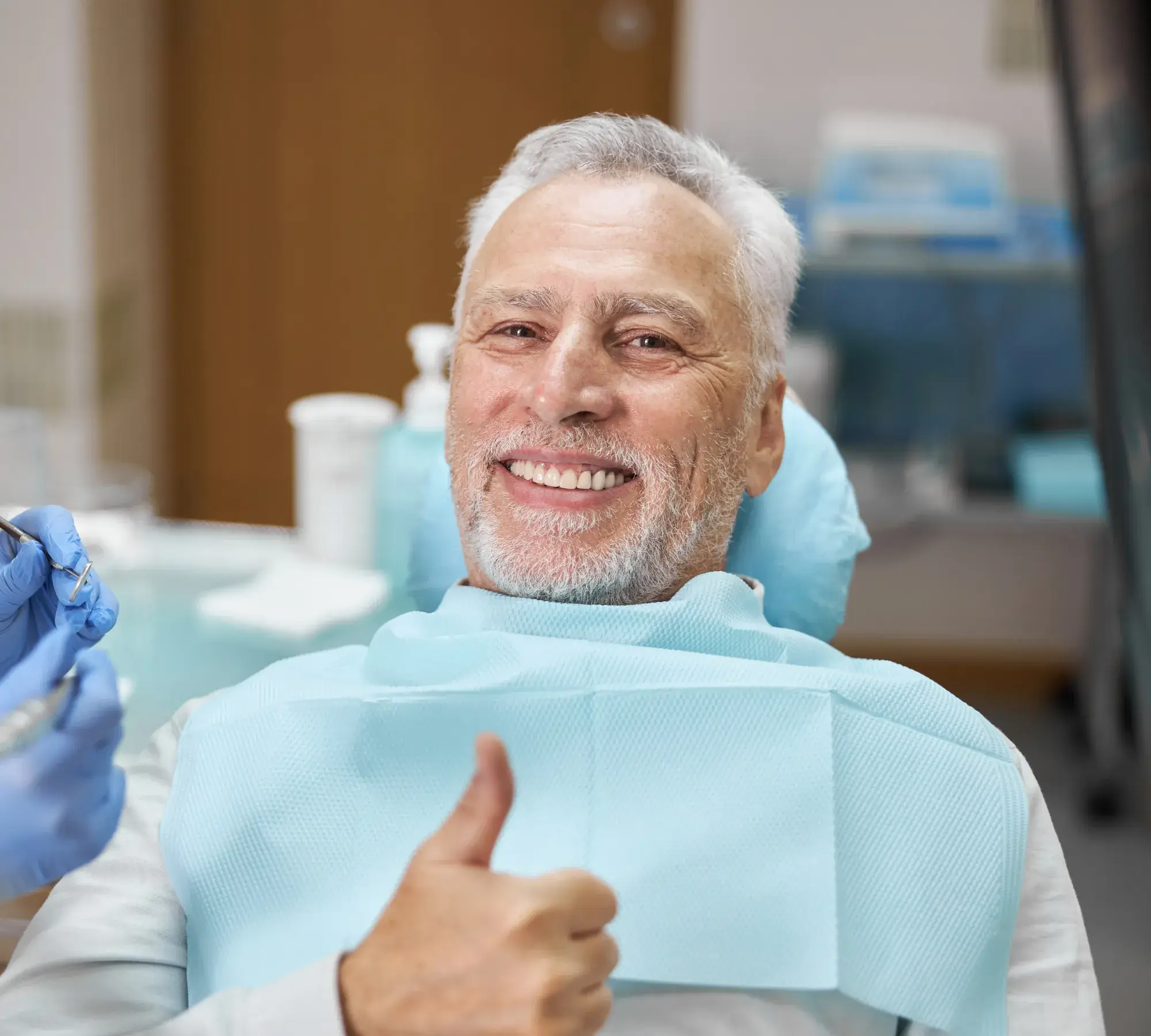 Smiling older man at dentist office giving thumbs up with dental bib and gloves visible.