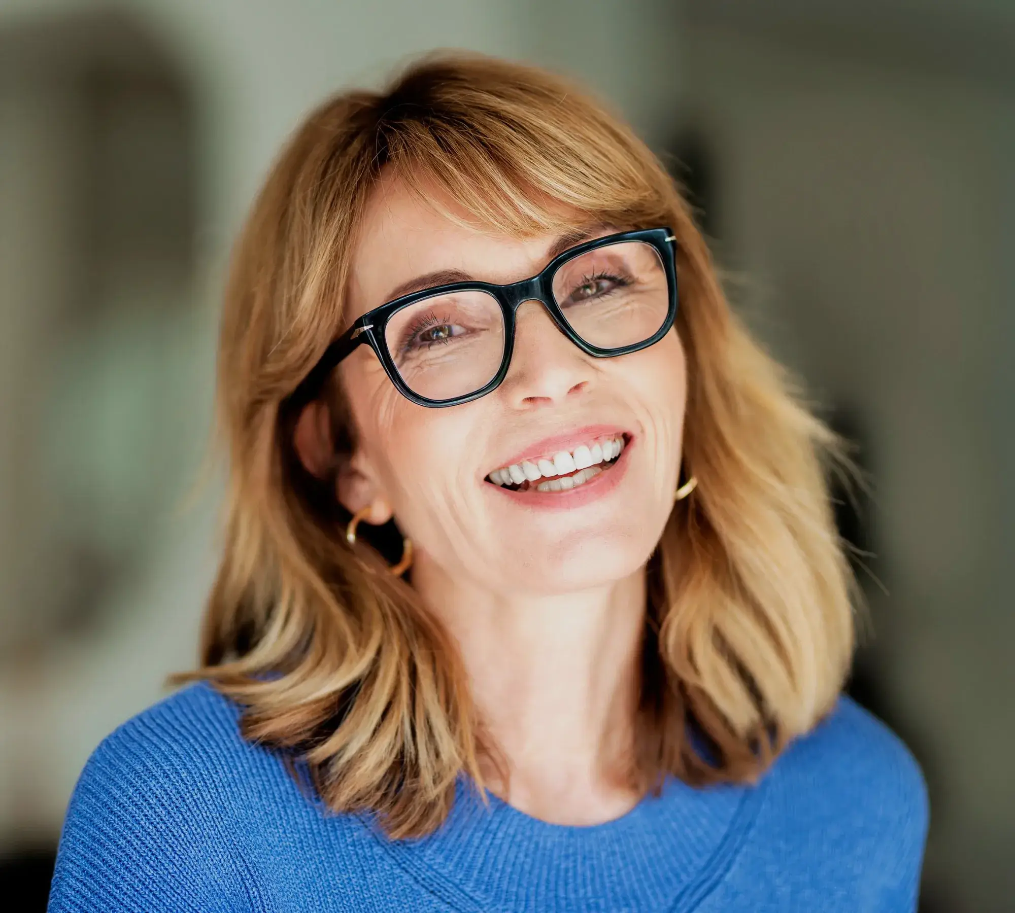 Smiling middle-aged woman with blonde hair, black glasses, gold hoop earrings, and blue sweater.