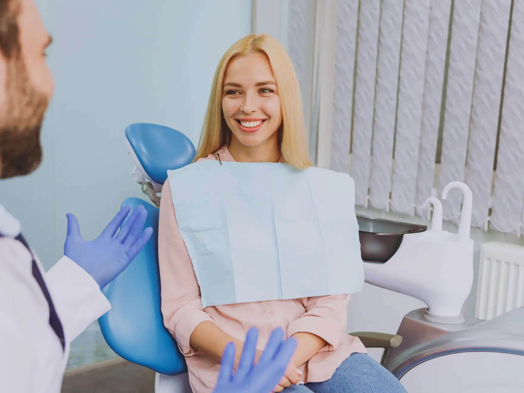 Smiling woman in a dental chair wearing a bib, talking to a dentist with blue gloves.