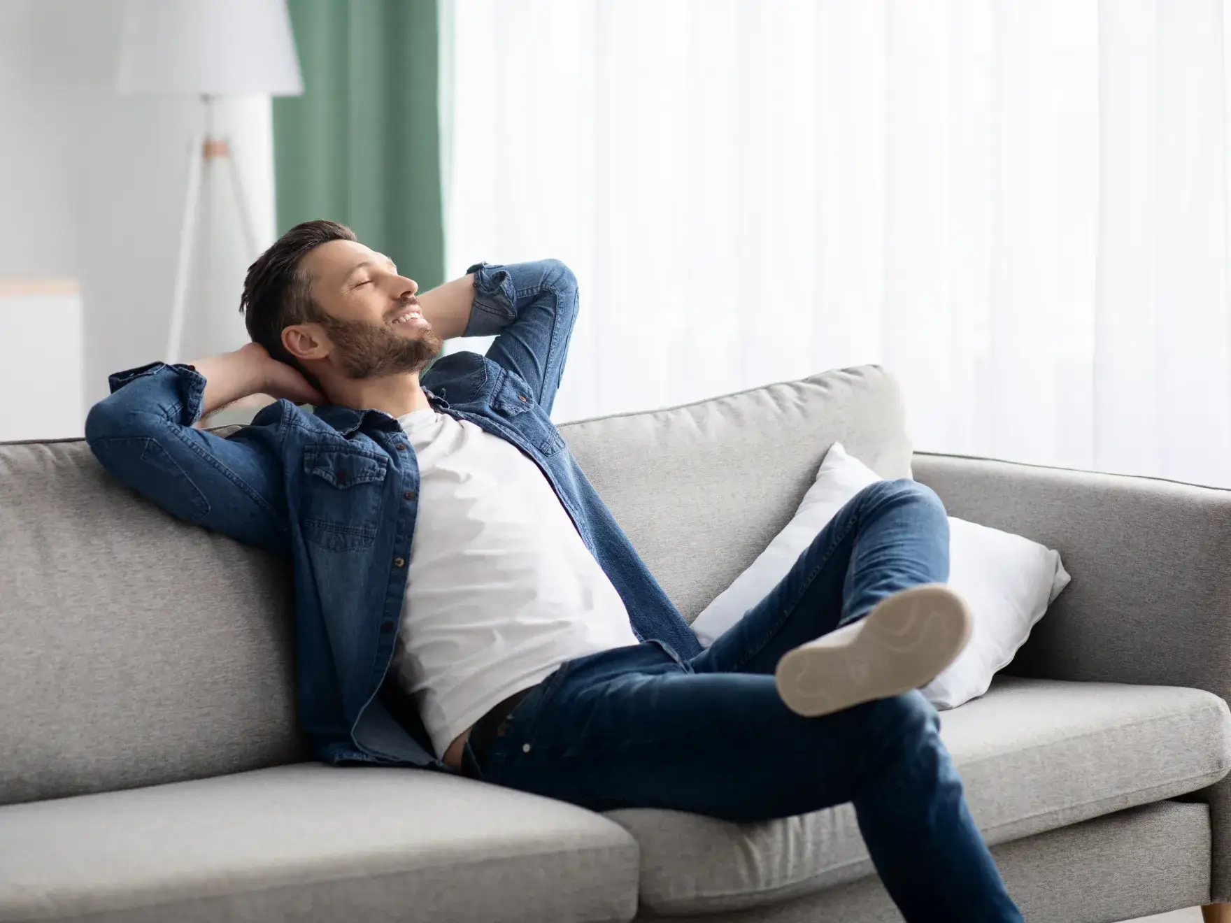 Man in denim jacket relaxing with eyes closed on a light gray couch by a window.