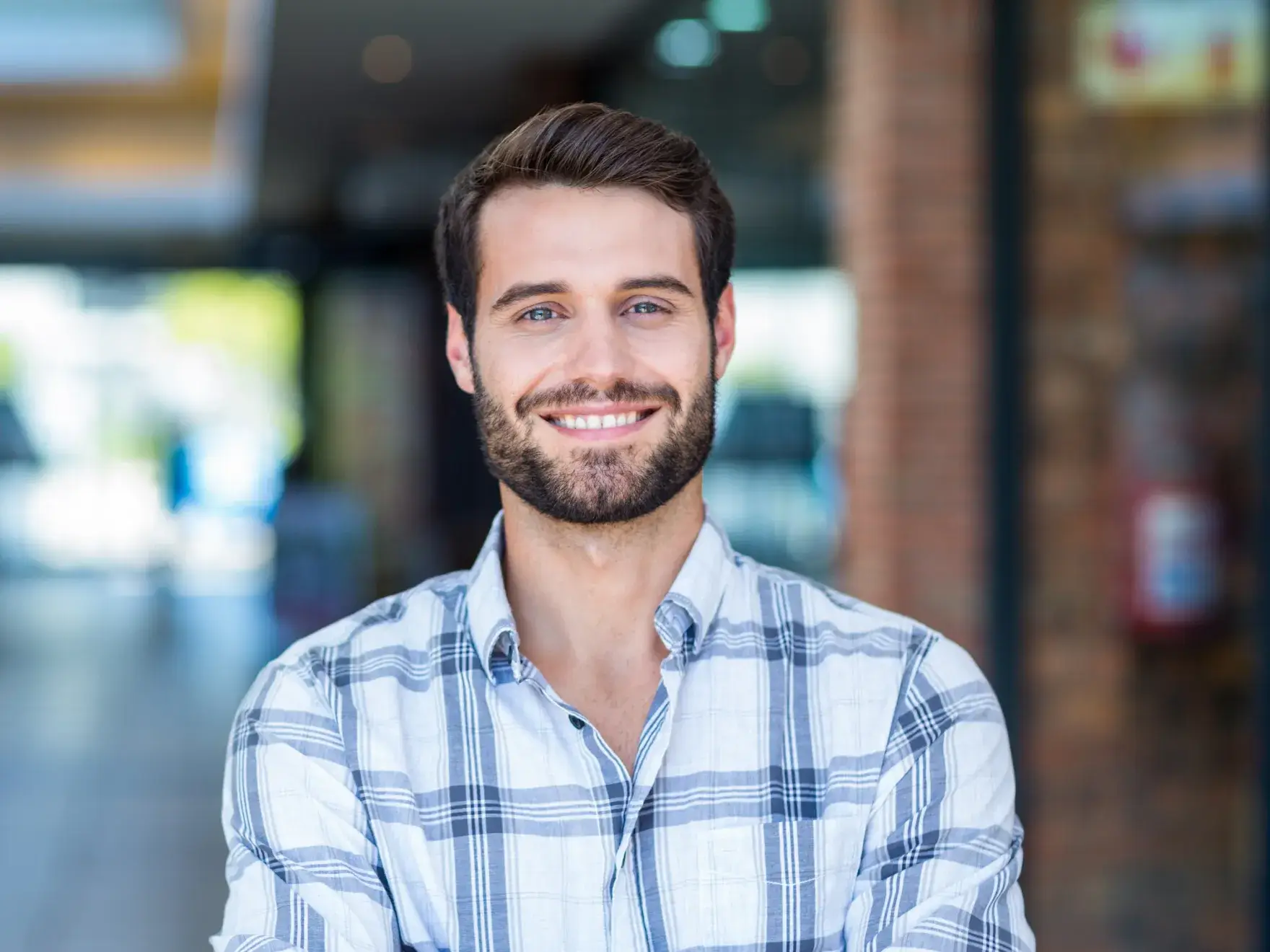 Smiling man with beard and plaid shirt standing indoors with blurred background.
