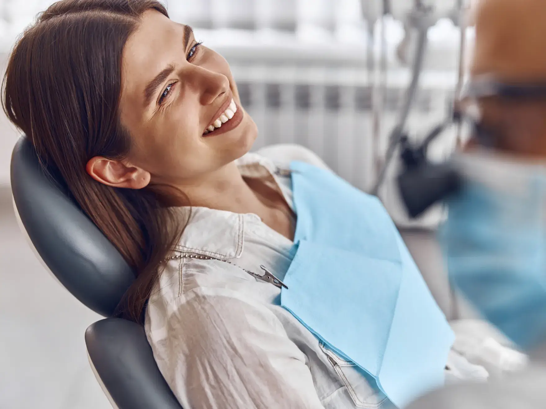Smiling woman reclined in dental chair wearing a blue bib, with dentist blurred in foreground.