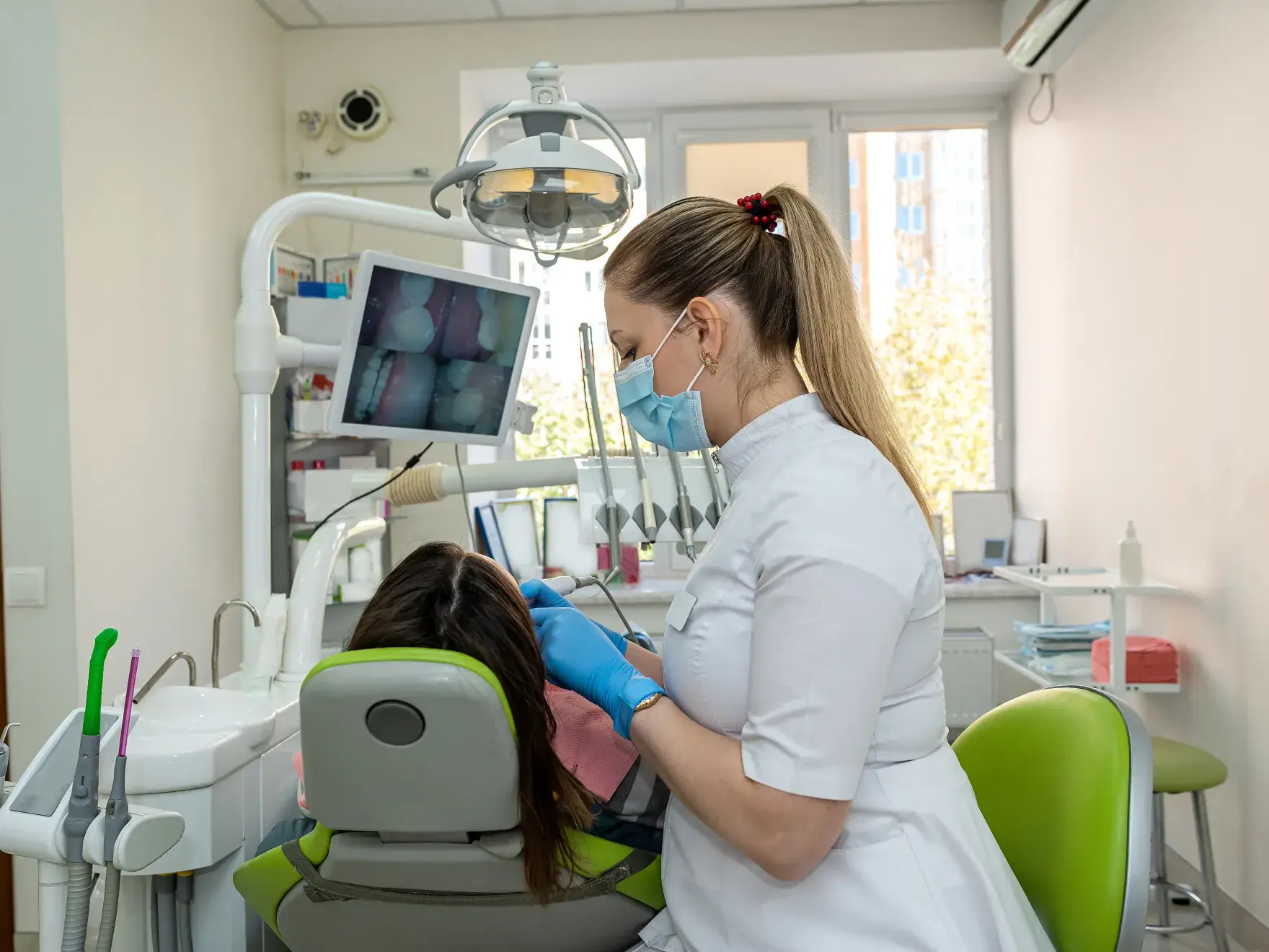Dentist wearing mask and gloves examining a patient in a dental clinic with dental X-rays on screen.