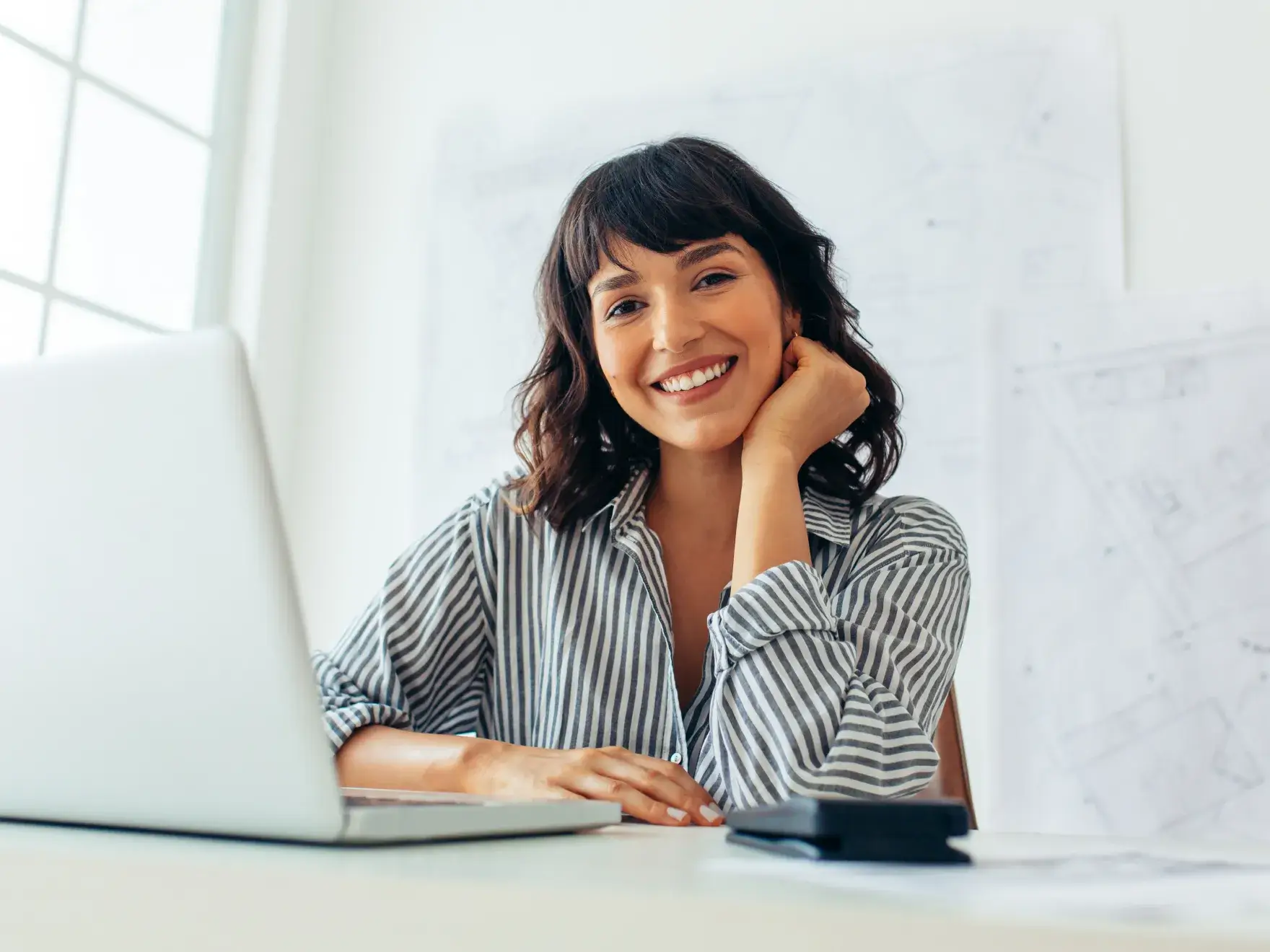 Smiling woman in striped shirt sitting at desk with laptop and phone in bright office.