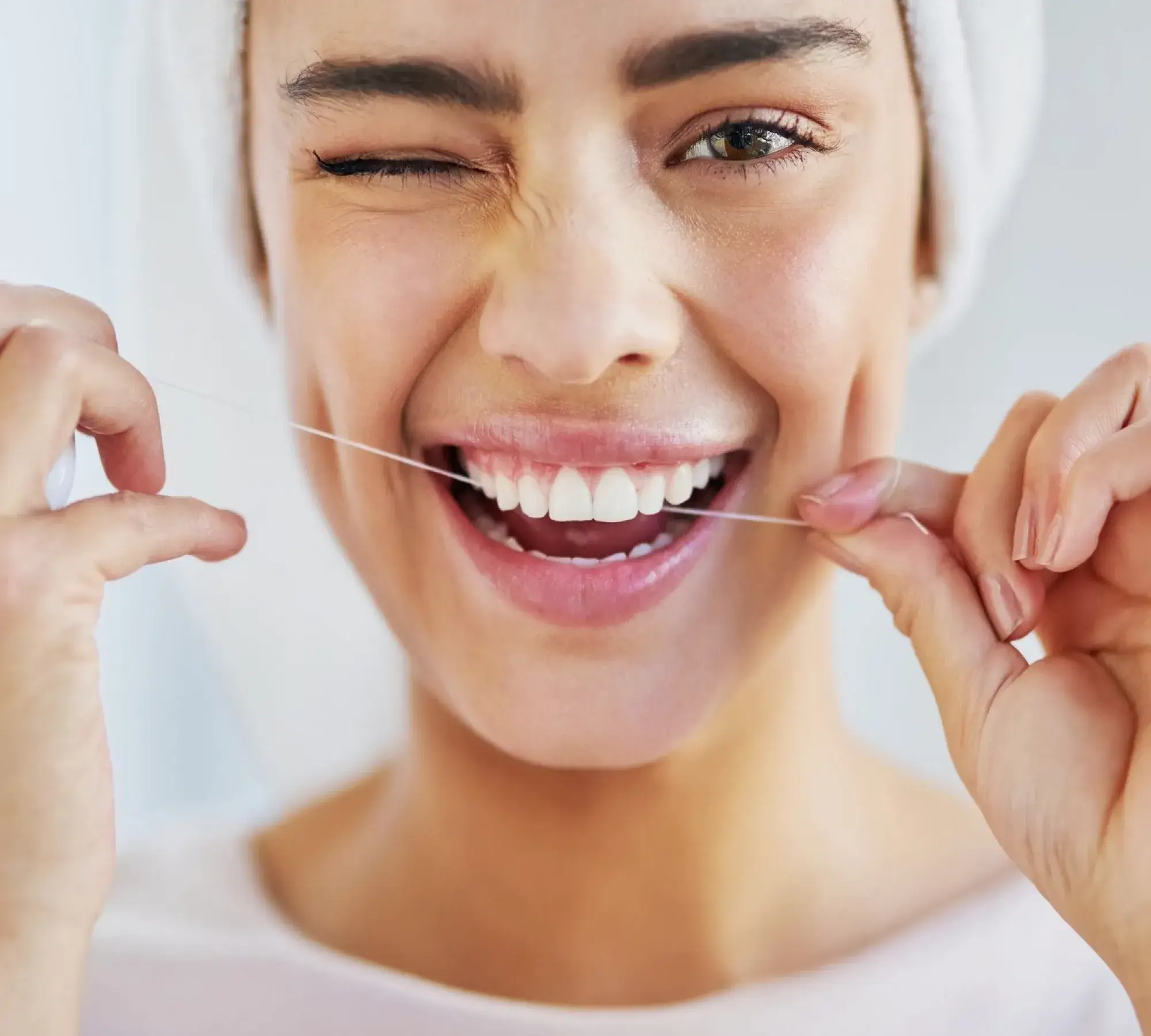 Close-up of a woman winking and smiling while flossing her teeth with dental floss.