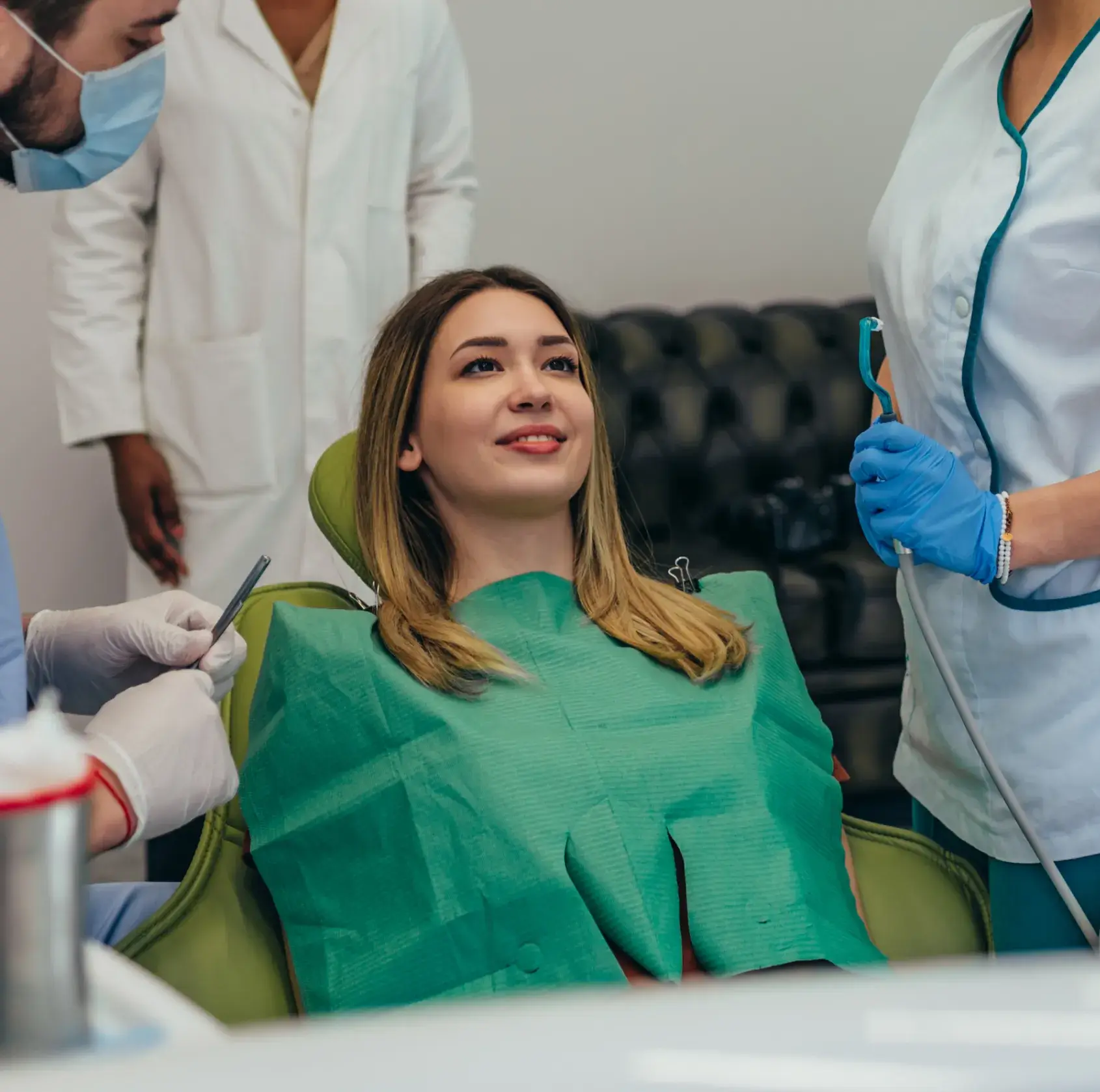 Smiling woman in dental chair with green bib, surrounded by dentists and dental tools.