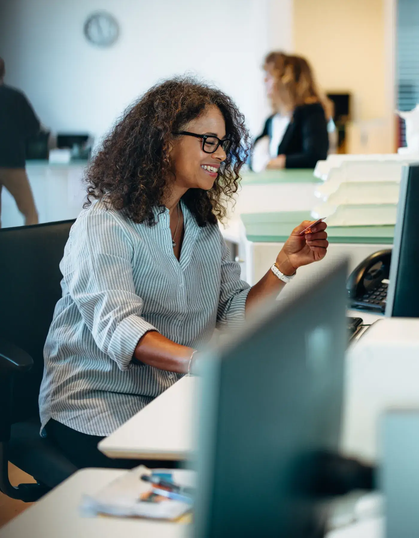 Smiling woman wearing glasses holding a card while sitting at an office desk with computer.