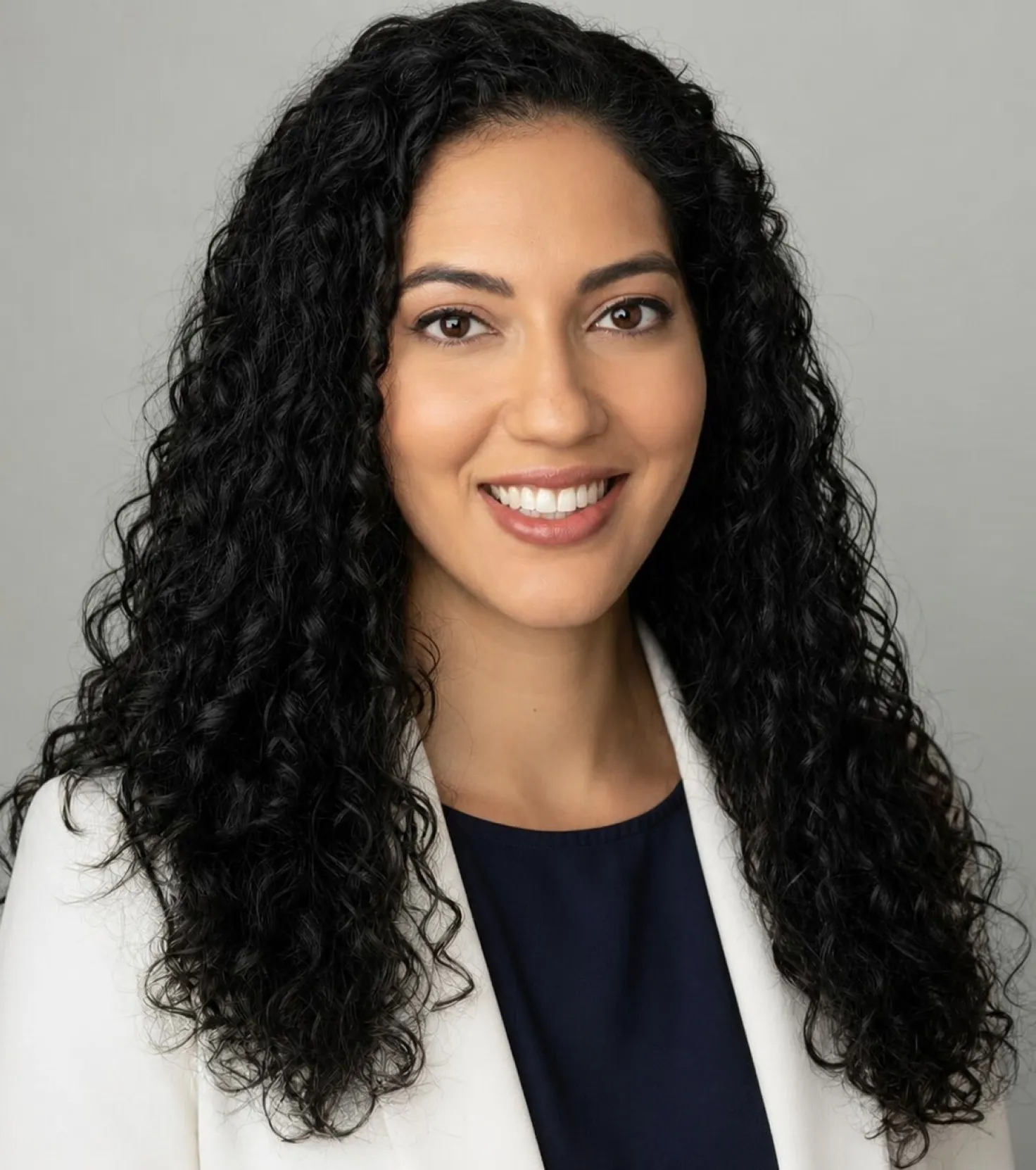 Smiling woman with long curly black hair wearing a white blazer and navy top against a gray background.
