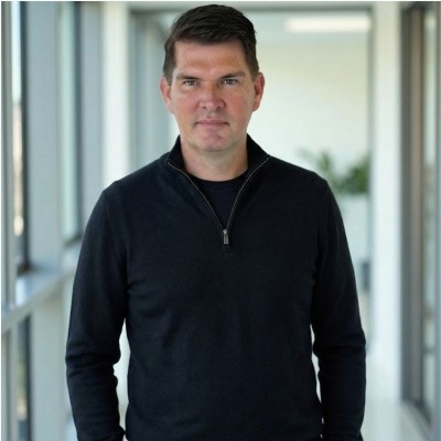 Man with dark hair wearing a black zip-up sweater standing in a bright hallway with windows.